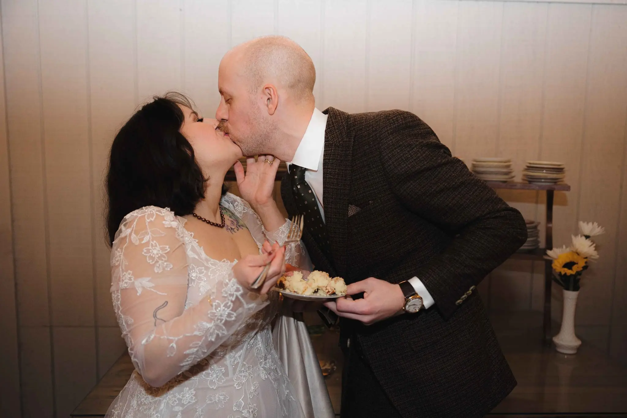 Couple kiss over a plate of wedding cake at the reception