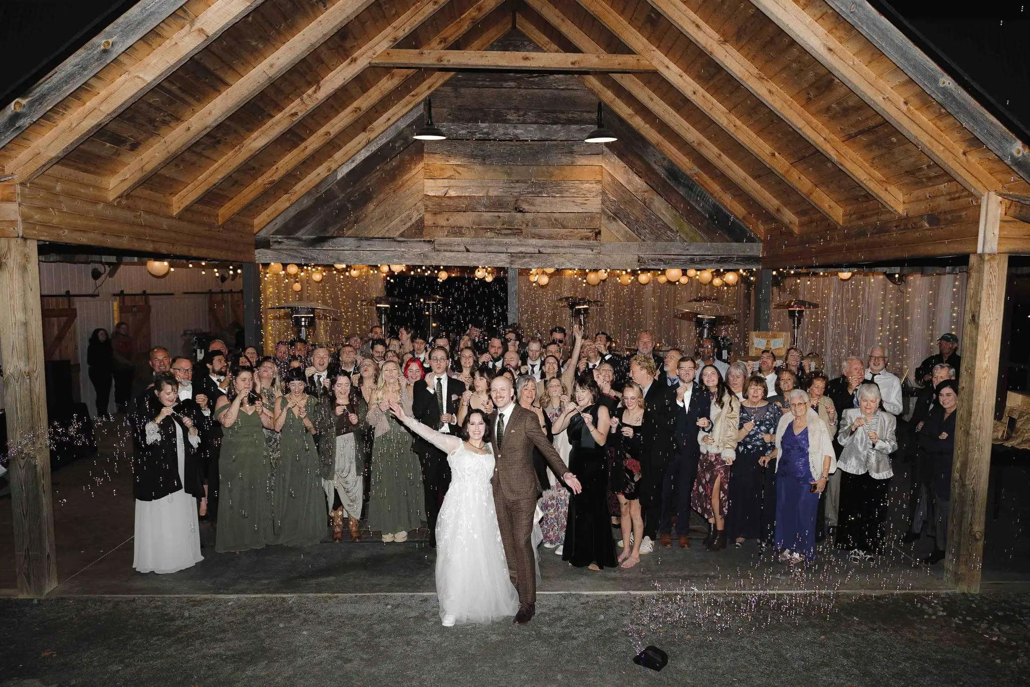 Newlyweds raise their arms as the whole wedding crowd cheers under the barn pavilion at night