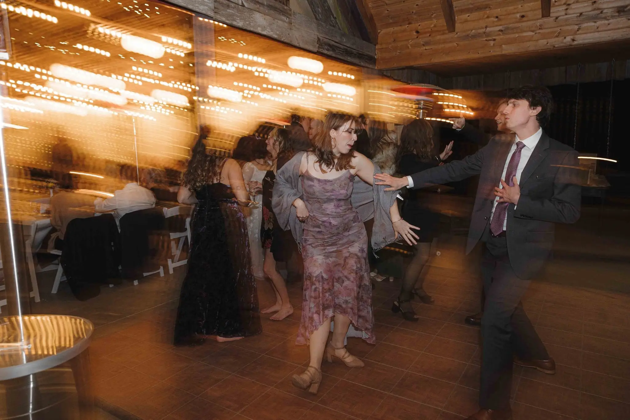 Motion blur of guests dancing under glowing string lights at the barn reception