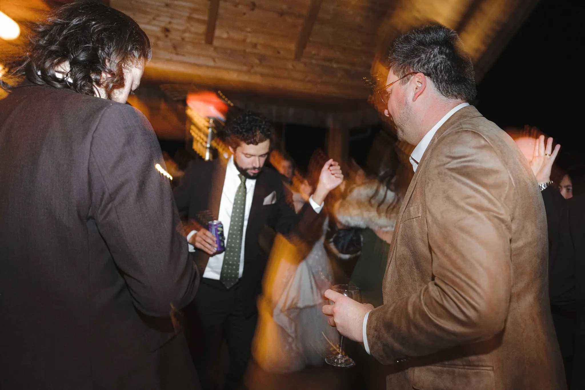 Groomsman in a green tie dances with a drink in hand as a guest in a tan jacket looks on