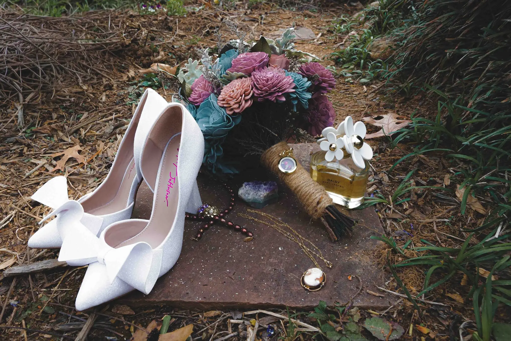 White bow heels, a wooden flower bouquet, perfume and jewelry arranged on a slab among fallen leaves