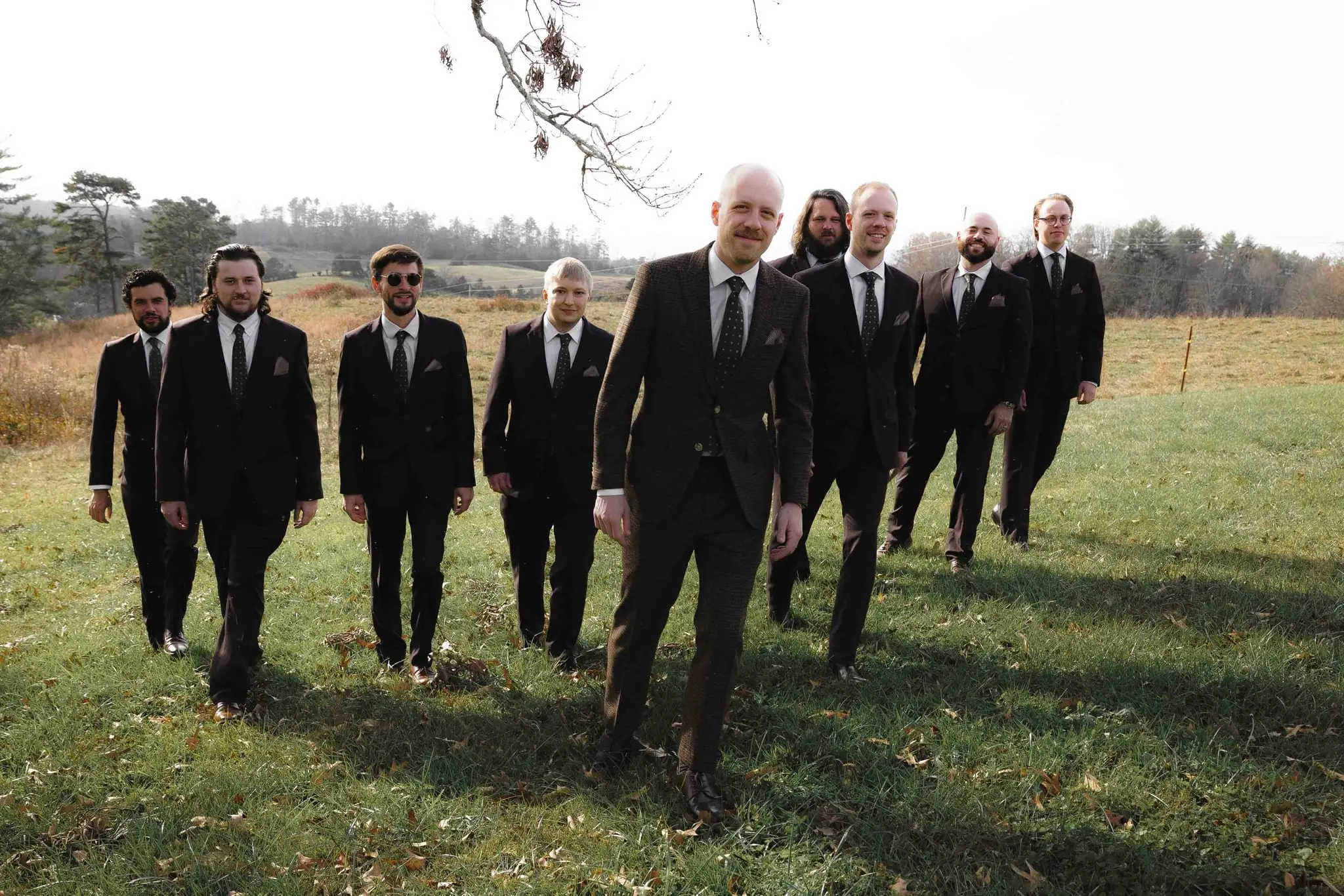 Groom and his groomsmen in dark suits walk toward the camera across a grassy pasture