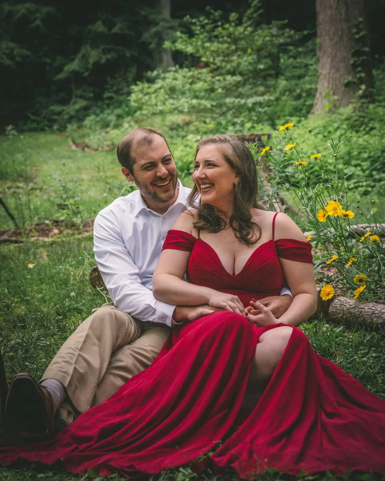 An engaged couple sitting in a wildflower garden, she in a long red dress leaning back against him