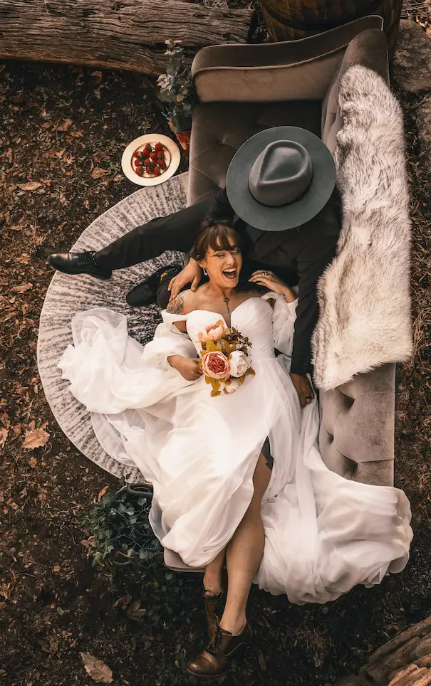 Overhead view of a couple lying on a rug among pillows and a fur throw, with a hat and fruit plate beside them