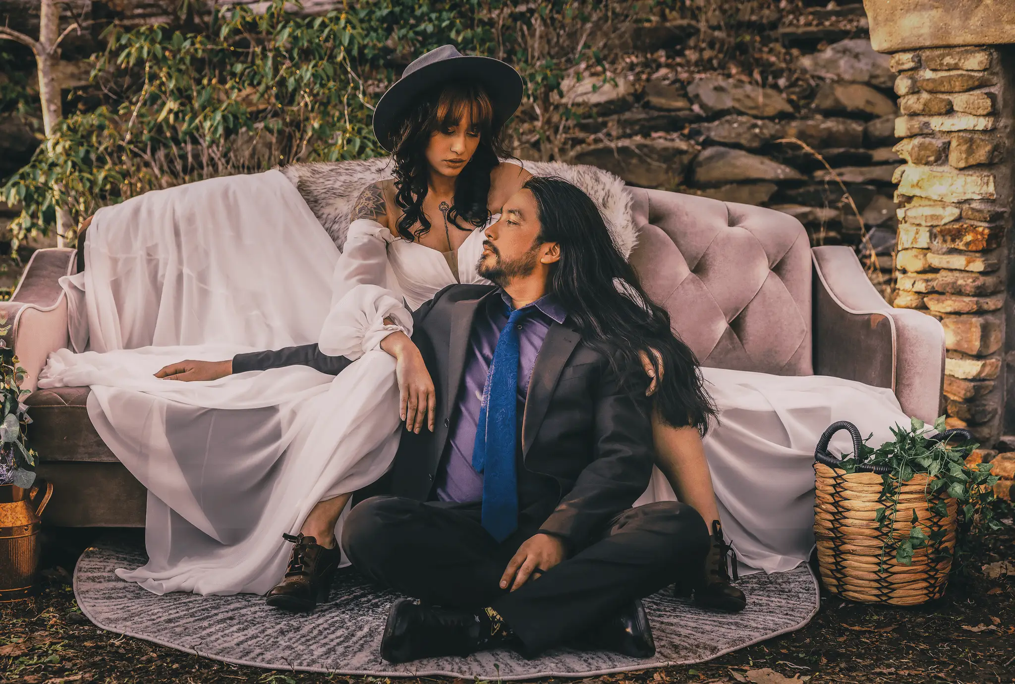 A couple posed on a pink vintage sofa outdoors, she in a wide hat, he seated on a rug in front
