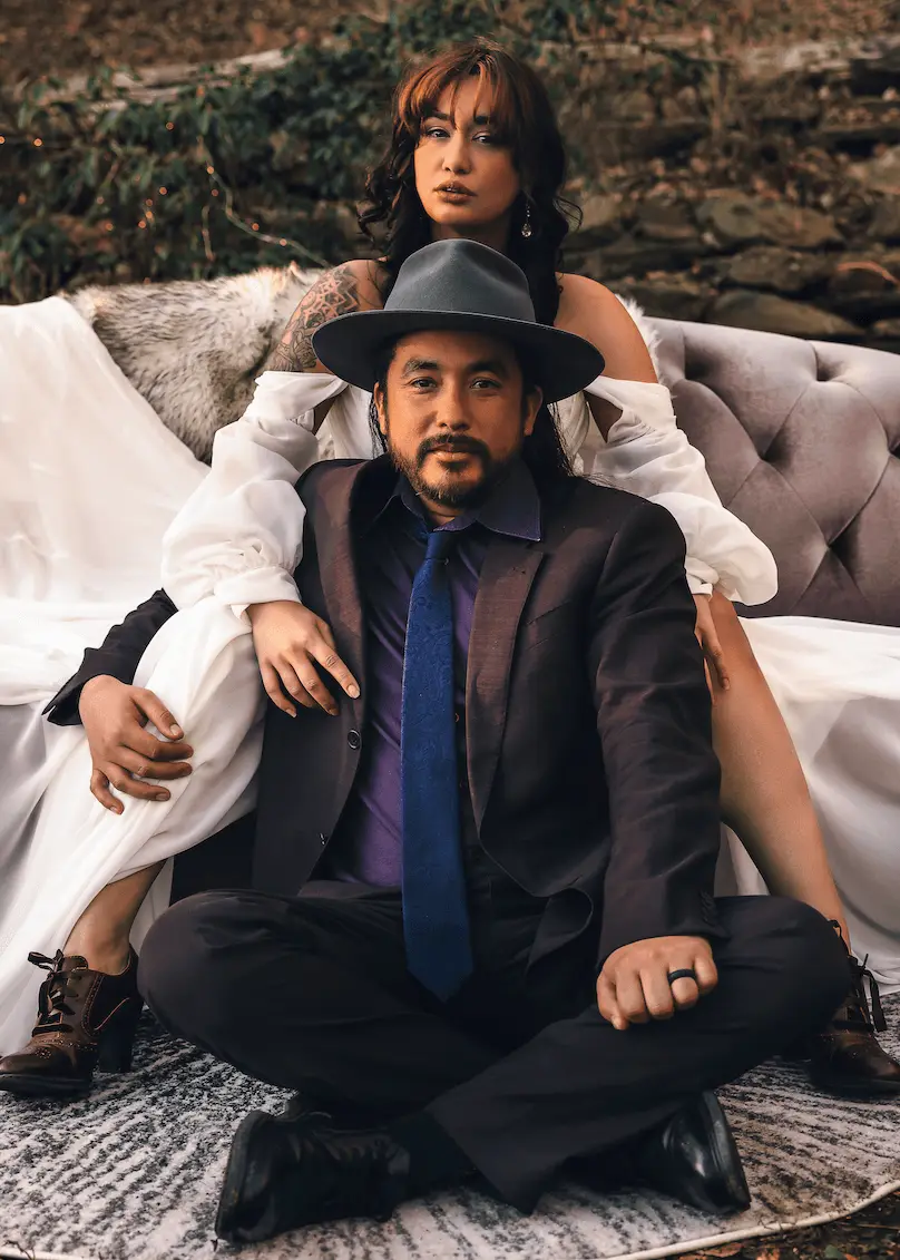 Juan and Jess posing on a mauve sofa, he seated cross-legged on a rug in a gray fedora