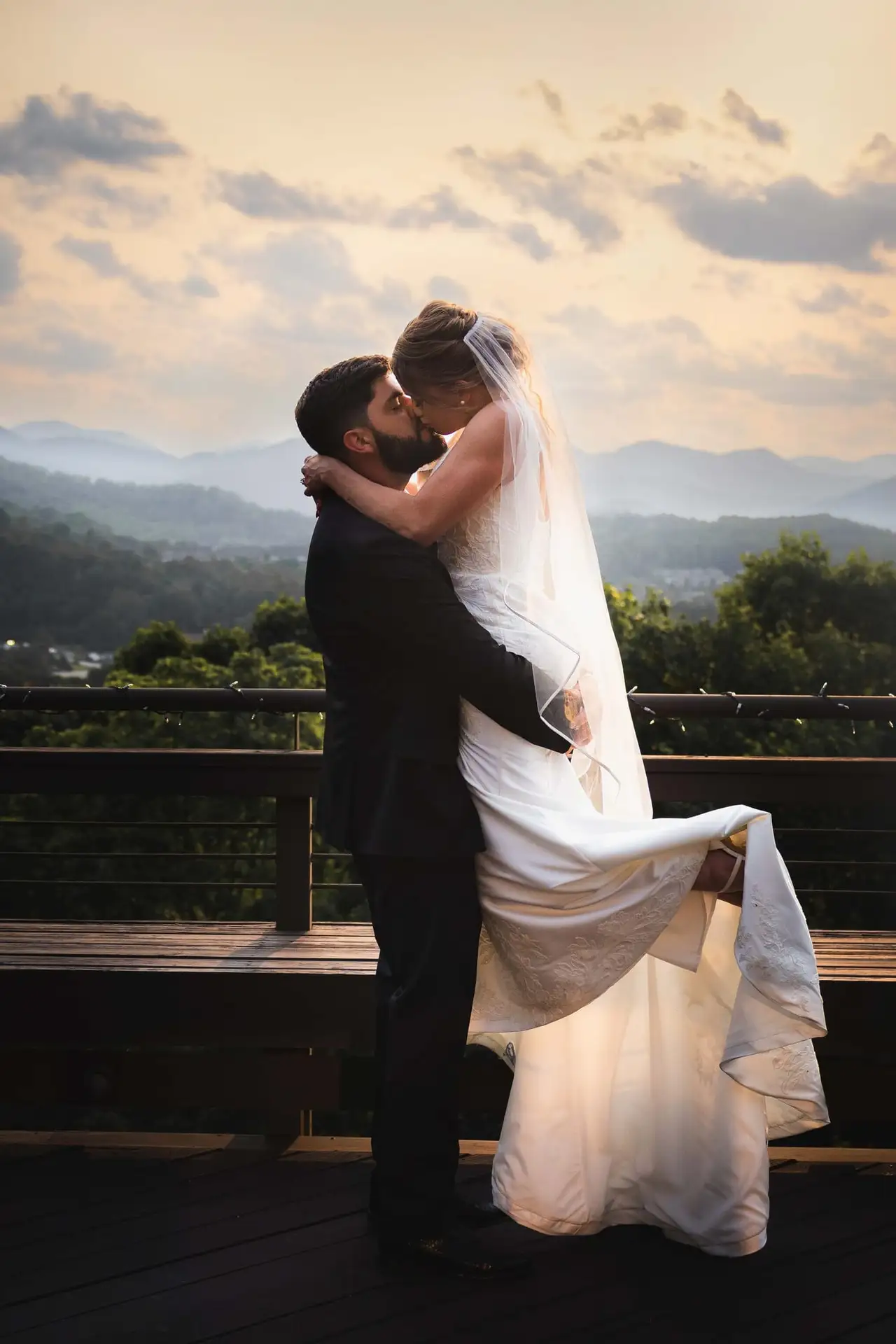 Groom lifting his bride for a kiss on a deck above the Blue Ridge mountains at sunset