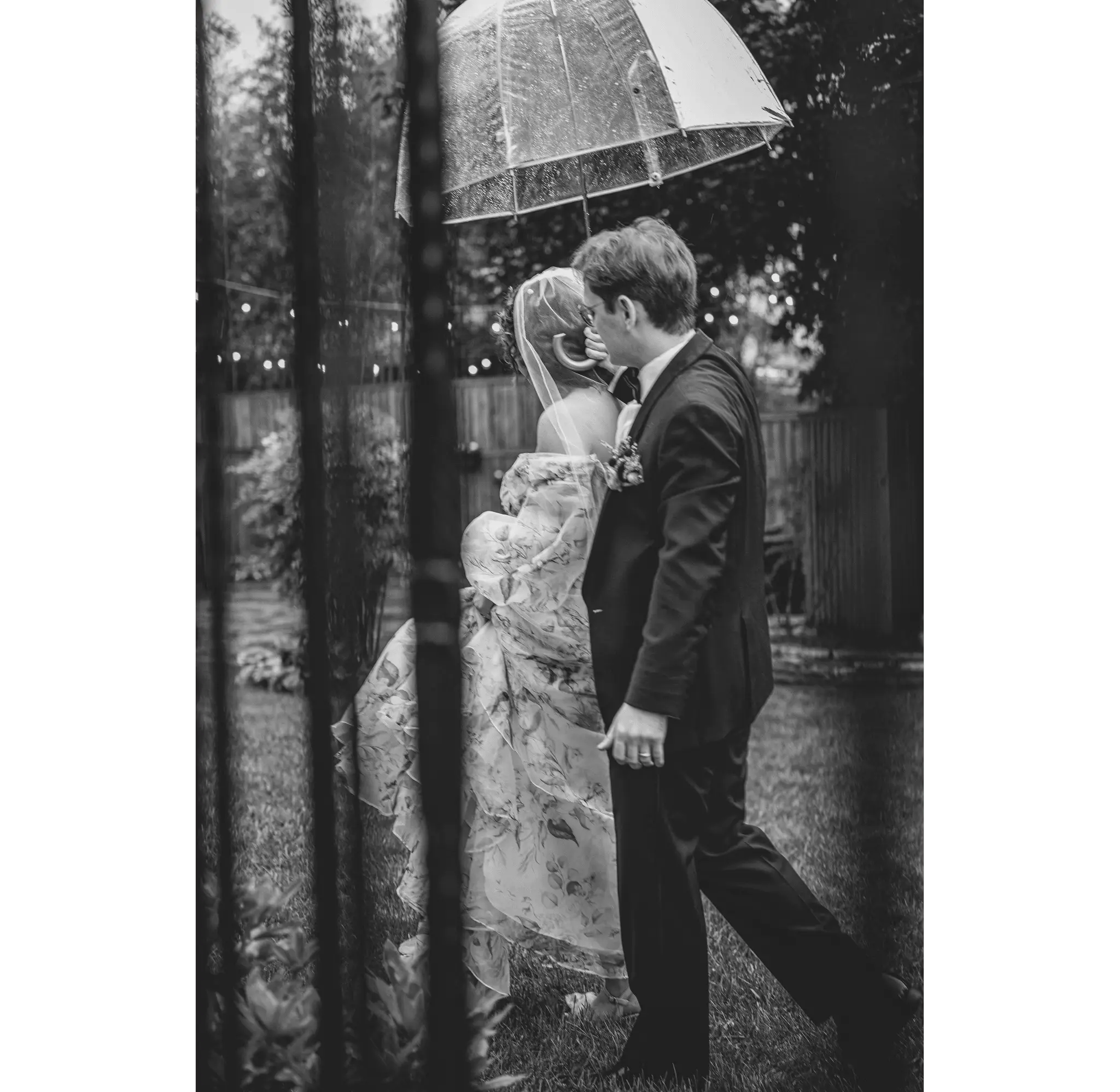 Black and white of the groom walking beside the bride under a clear umbrella, string lights behind them