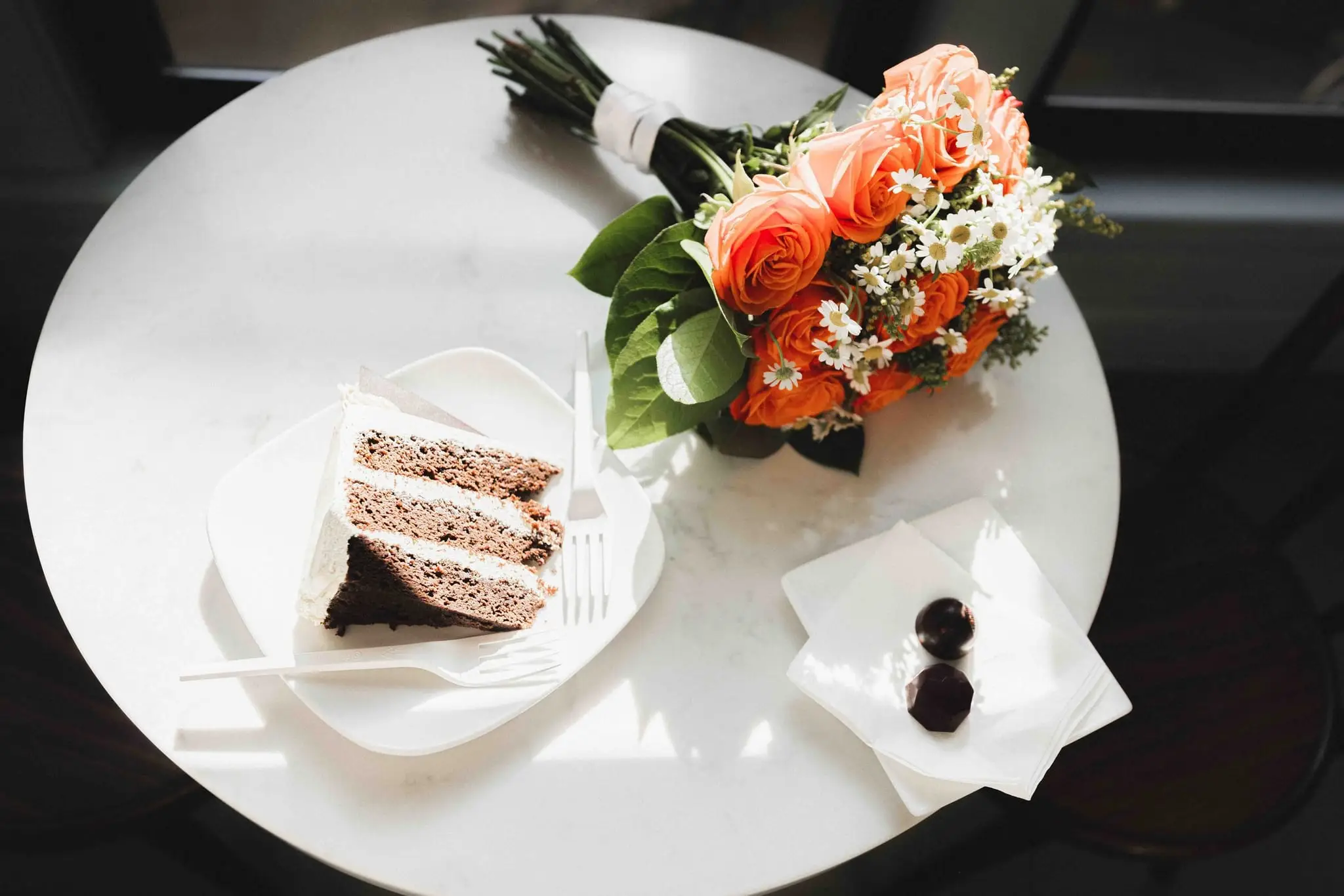 Overhead view of a slice of wedding cake, a bouquet of orange roses and two chocolates on a white cafe table