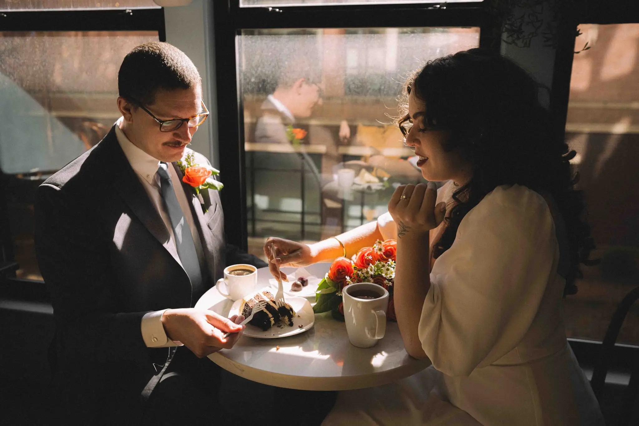Giuliette and Will’s courthouse wedding day, Asheville NC — the groom reaching toward cake and coffee on a cafe table as the bride laughs