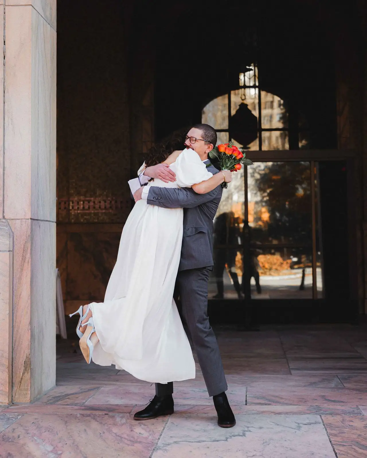 Giuliette and Will wedding at Buncombe County Courthouse, Asheville NC — the groom lifting the bride in a hug beneath a stone archway, her bouquet in hand