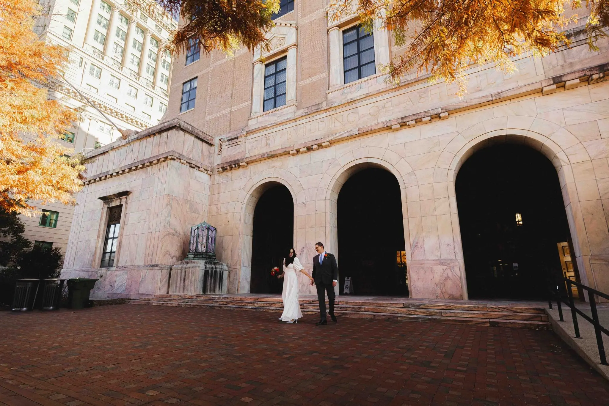 Giuliette and Will wedding at Buncombe County Courthouse, Asheville NC — the couple small in front of a stone building with three arched doorways and orange autumn leaves