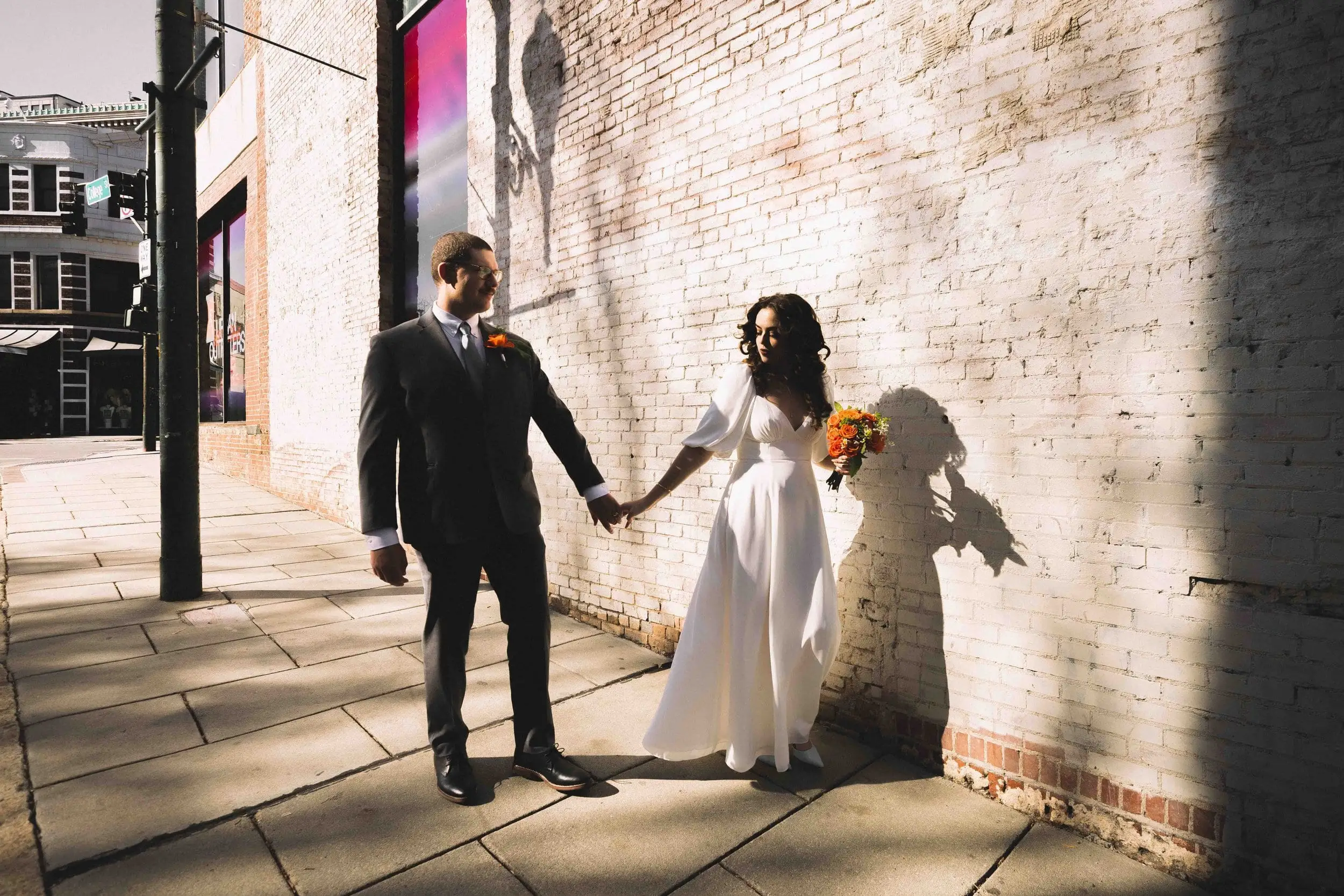 Giuliette and Will’s courthouse wedding day, Asheville NC — the couple holding hands beside a brick wall in long afternoon shadows