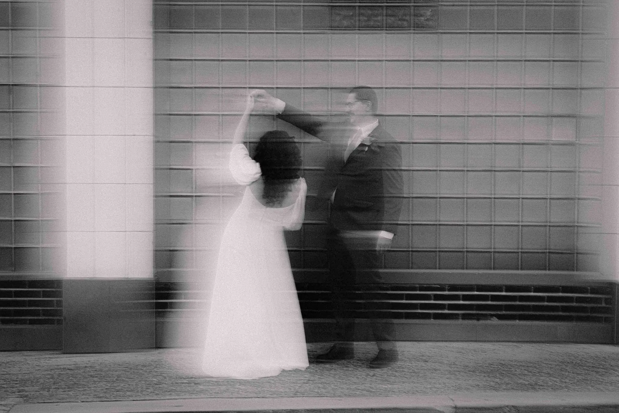 Black and white motion blur of a bride and groom twirling in front of a glass-block wall