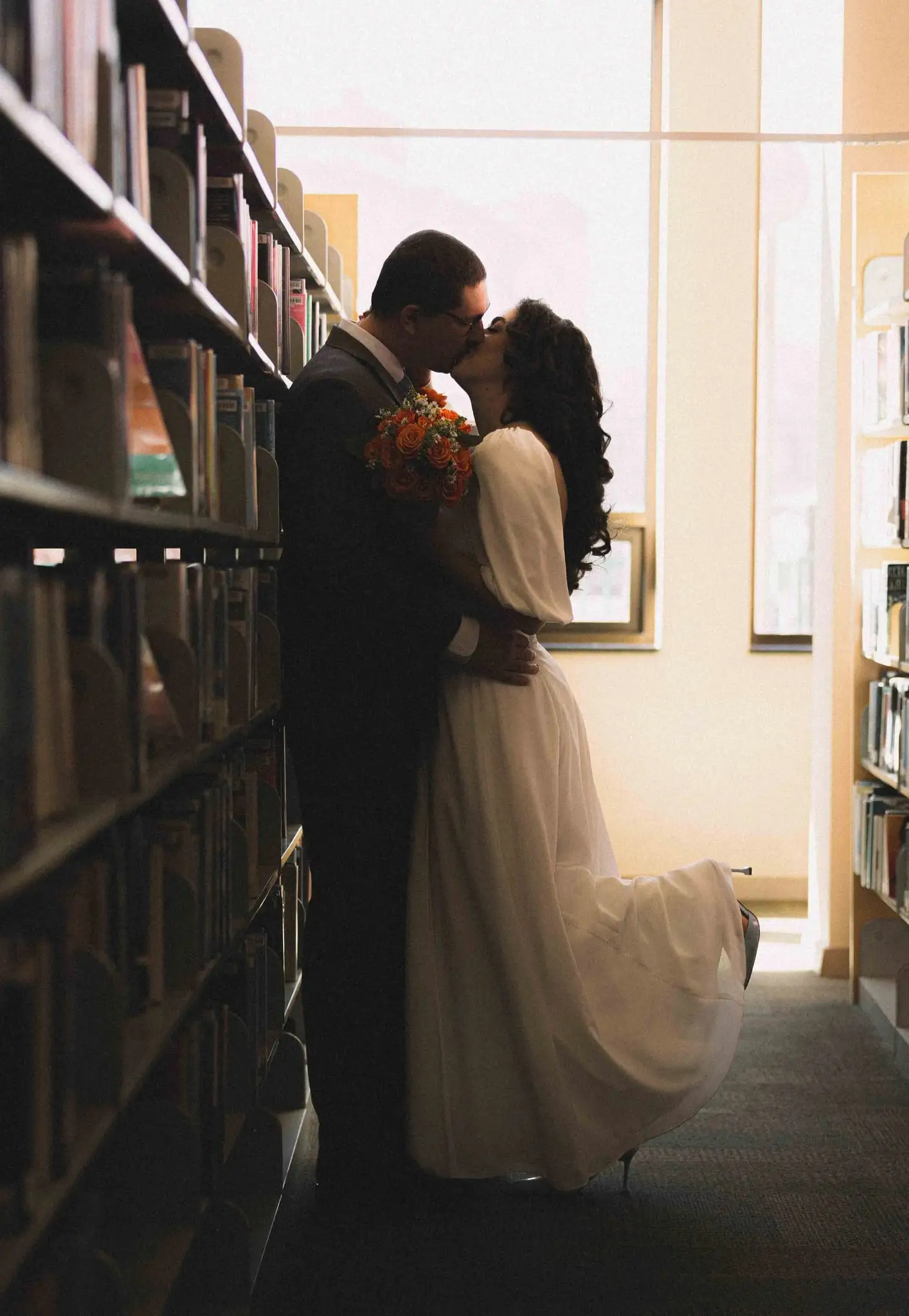 Couple kissing between library bookshelves in warm window light, the bride's skirt swaying