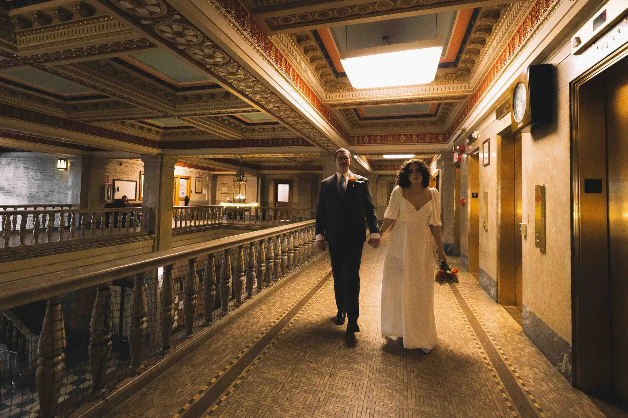 Giuliette and Will’s courthouse wedding day, Asheville NC — the couple walking along an ornate corridor with a railing and warm light