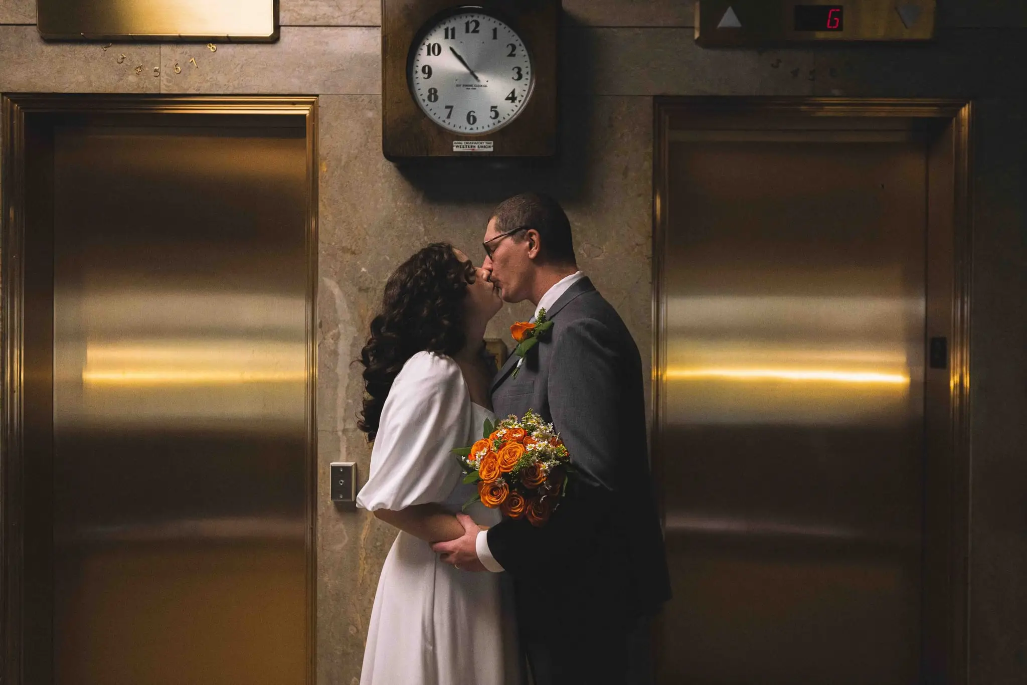 Giuliette and Will’s courthouse wedding day, Asheville NC — the couple kissing beside brass elevator doors and a wall clock