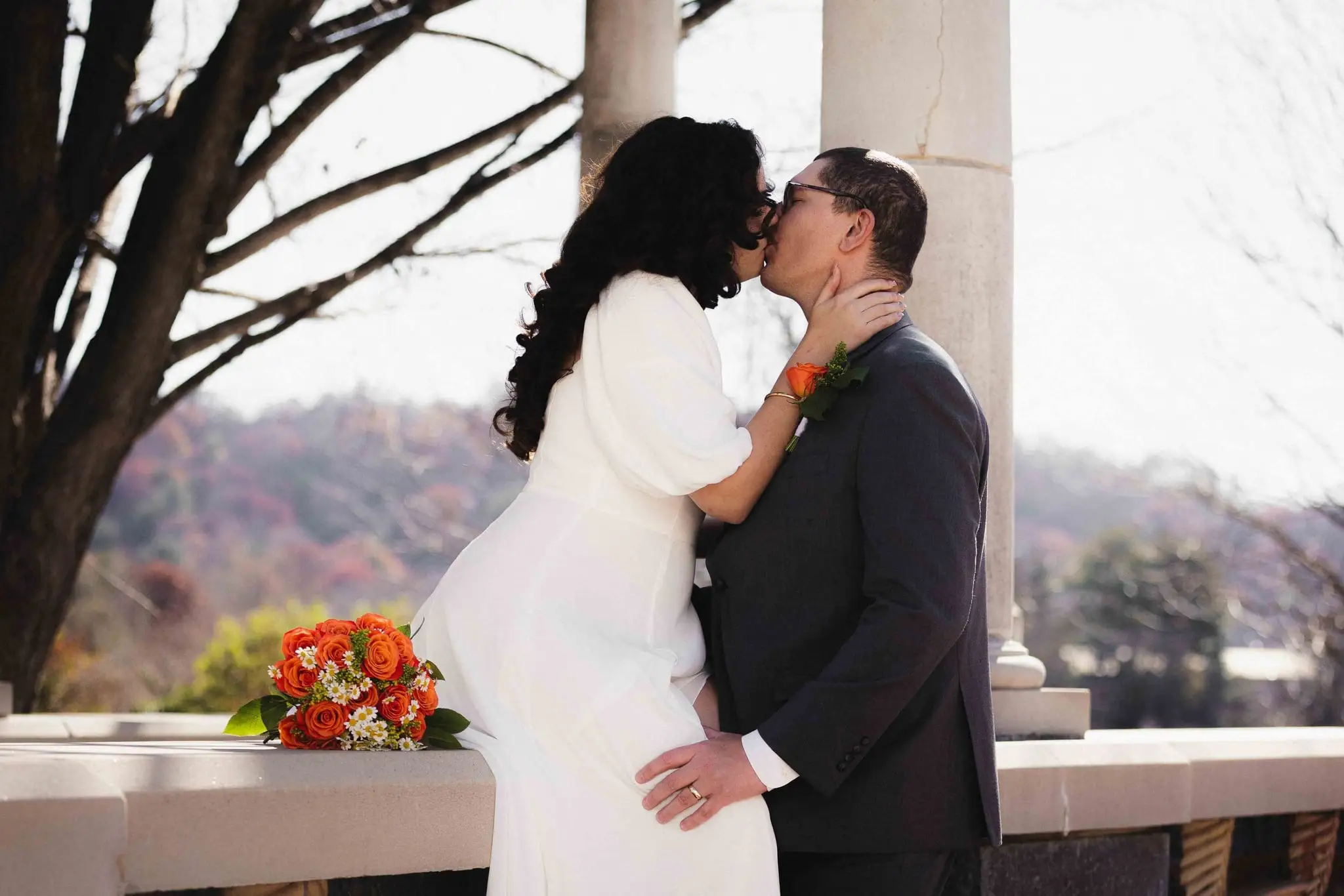 Giuliette and Will’s courthouse wedding day, Asheville NC — the couple kissing at a stone terrace railing with hills in the distance, an orange bouquet in the bride's hand
