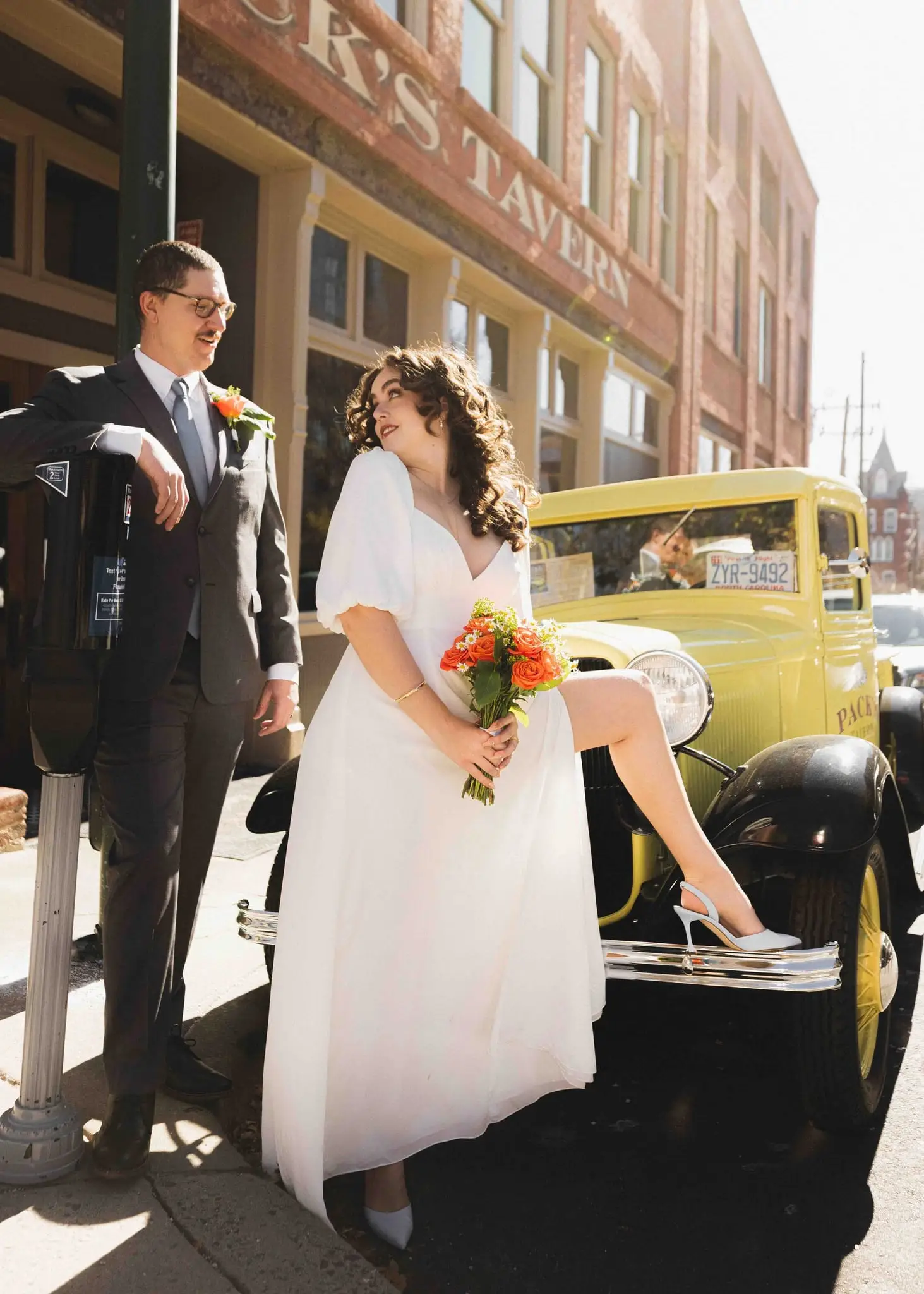 Giuliette and Will’s courthouse wedding day, Asheville NC — the bride seated on a yellow vintage car in front of a brick building, the groom beside her