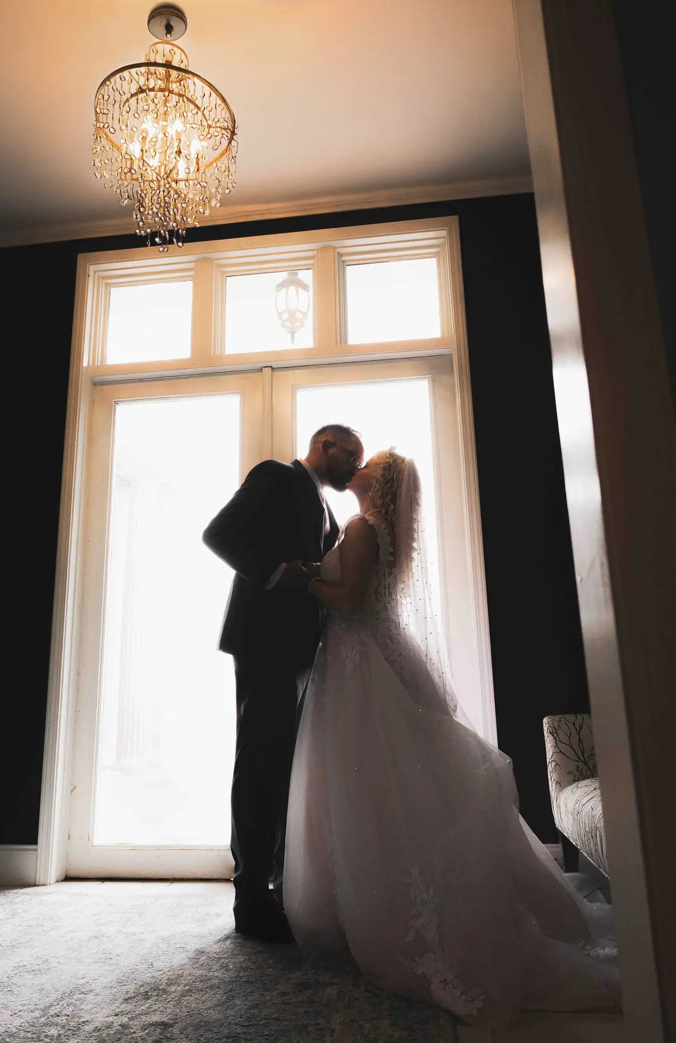 Couple kissing in silhouette against tall bright windows under a crystal chandelier