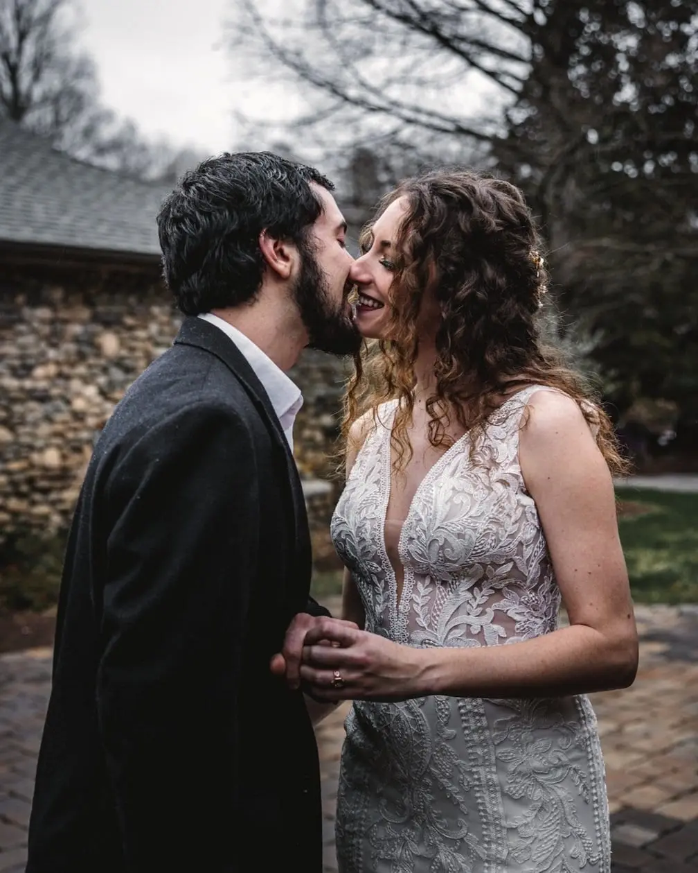 A couple smiling and touching noses outdoors, dressed in wedding attire, with trees and a stone building in the background.