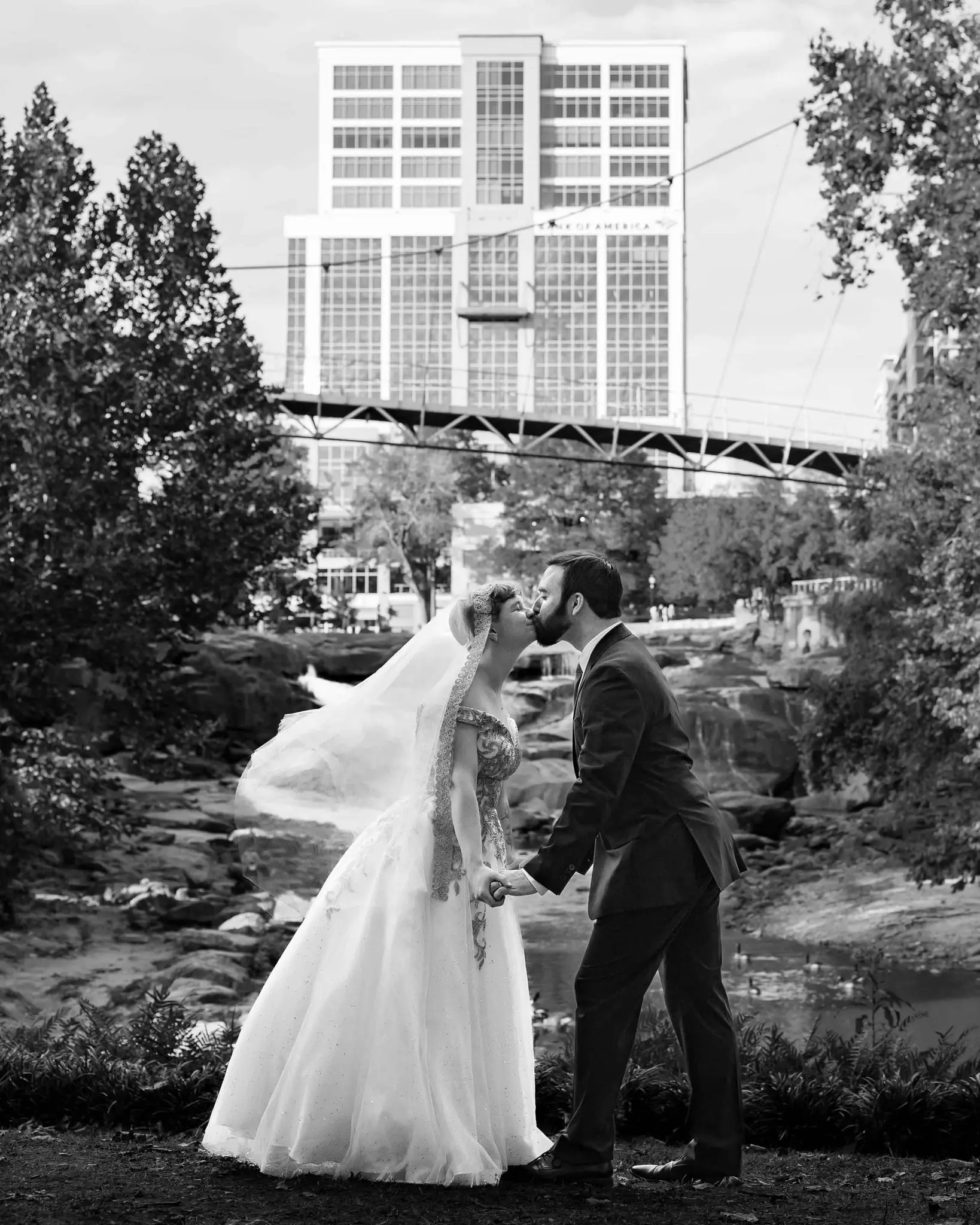 Black and white kiss as the bride's veil blows, with the river and a Greenville building behind the couple