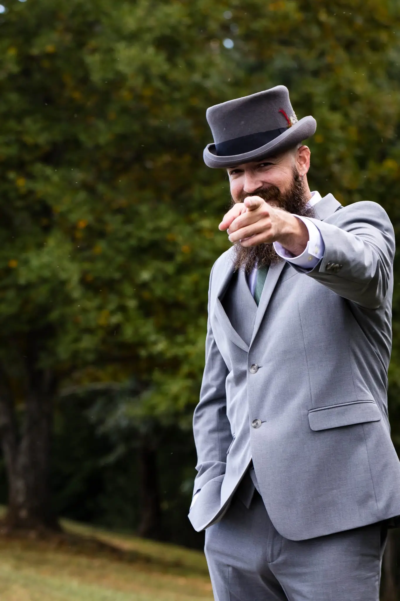 Groom in a gray suit and top hat points playfully at the camera in the woods