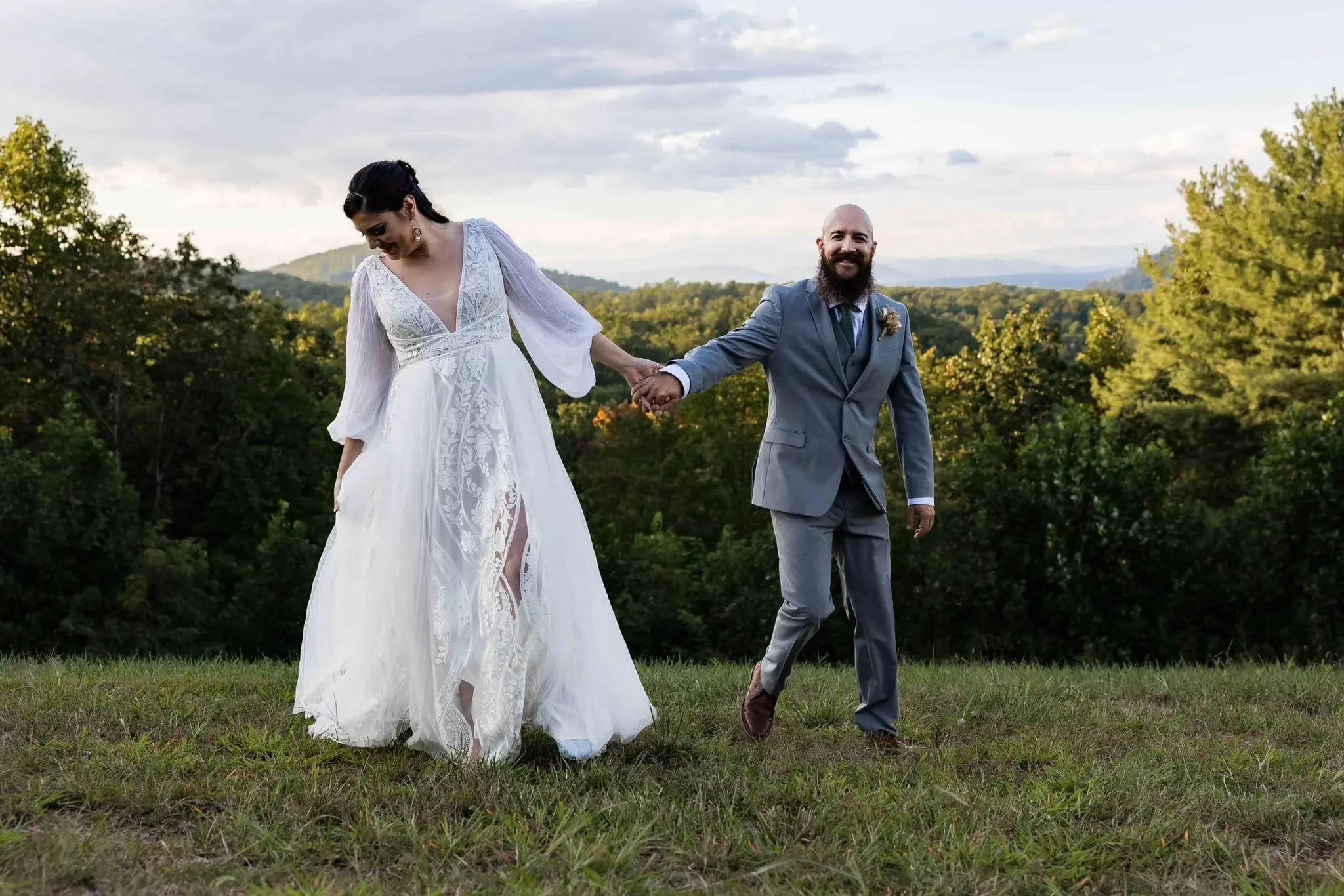 Bride walking in step with the groom, holding hands on the grassy overlook at sunset