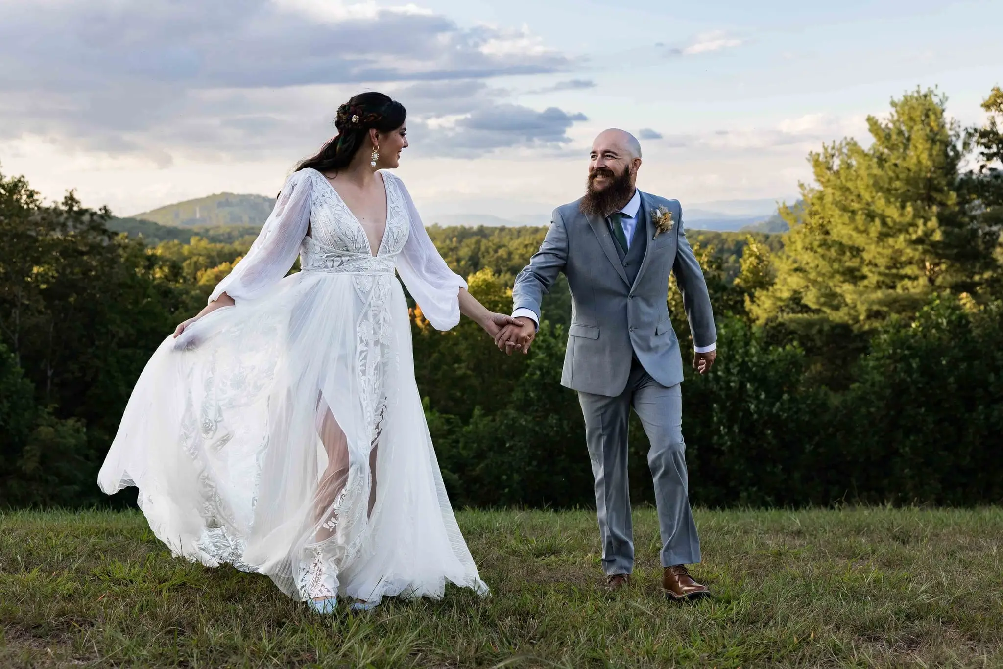 Bride and groom holding hands and walking across a hilltop lawn toward distant mountain ridges