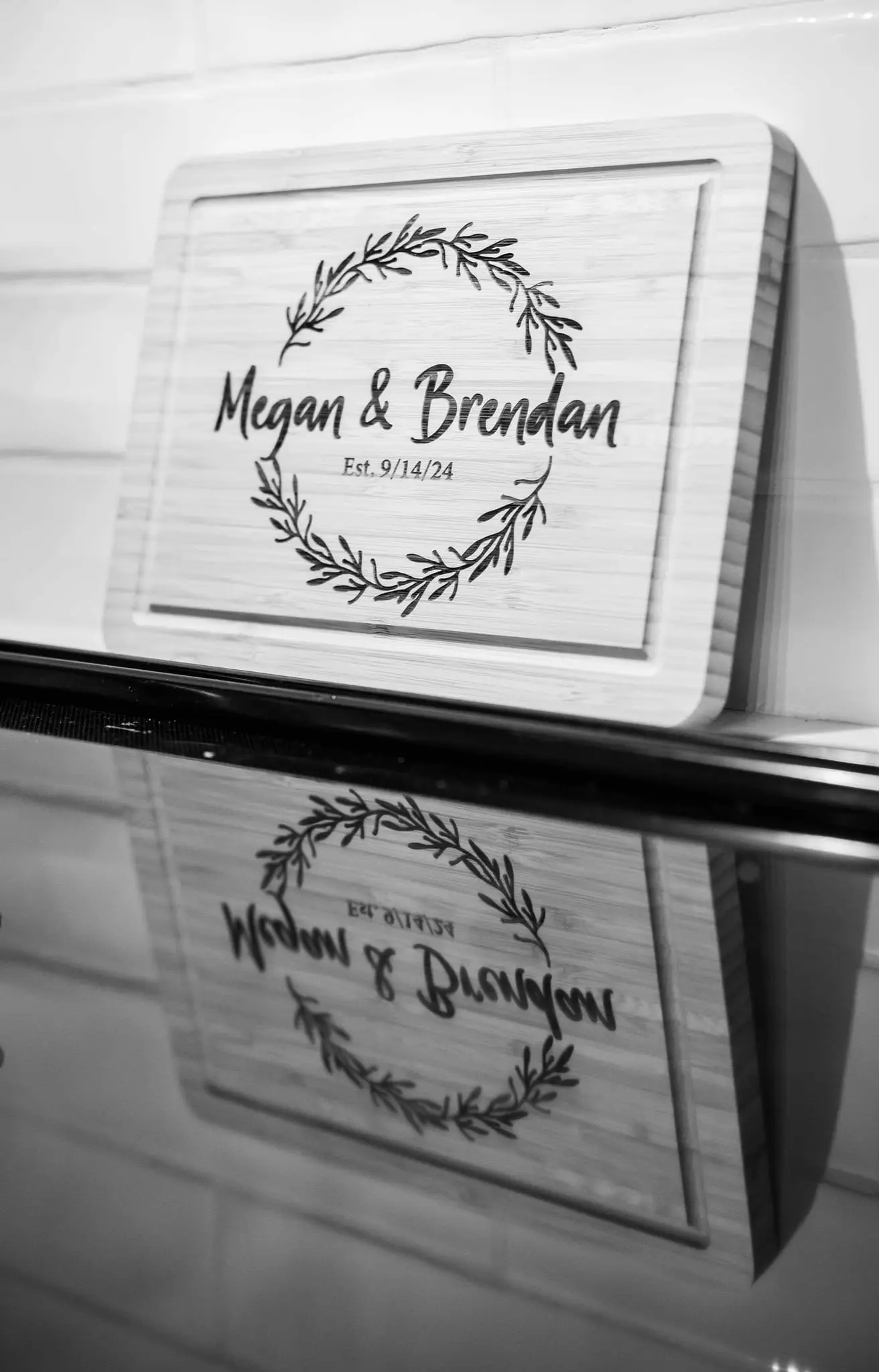 Engraved wooden cutting board reading Megan & Brendan inside a laurel wreath, reflected on a black counter