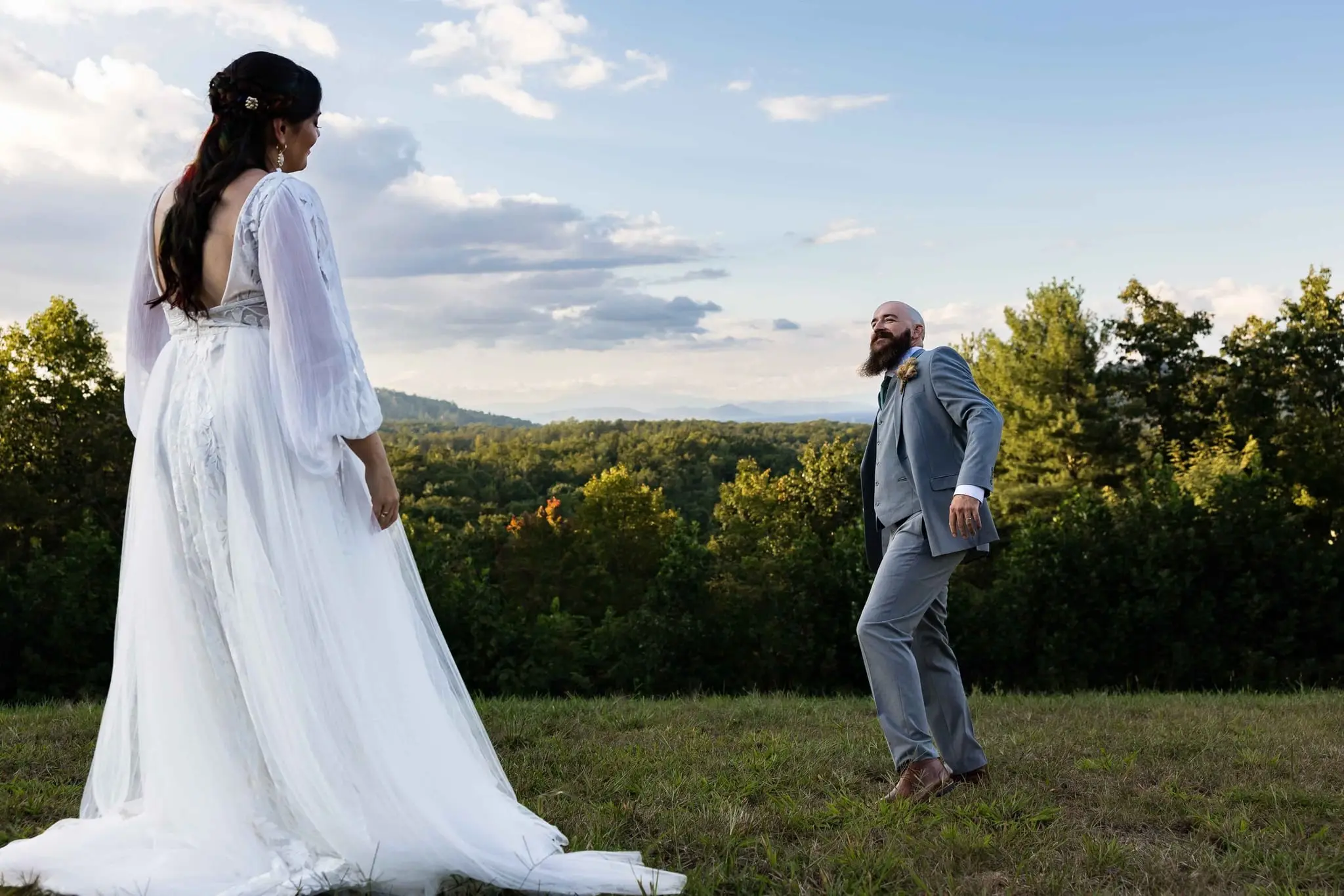 Bride watching the groom stroll away across the grassy hilltop, forested ridges beyond