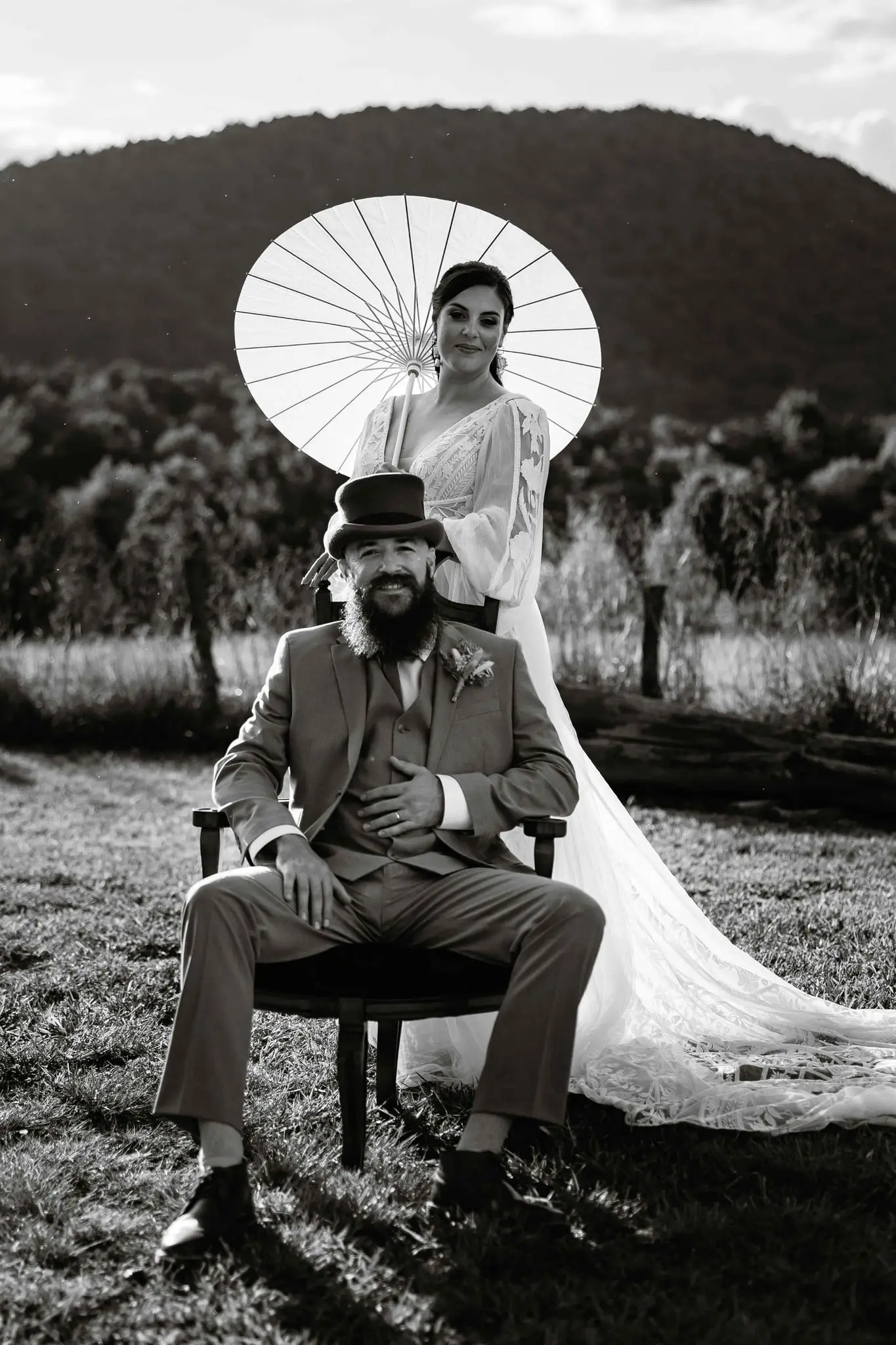 Black and white portrait of the groom seated in a bentwood chair beneath the bride's paper parasol