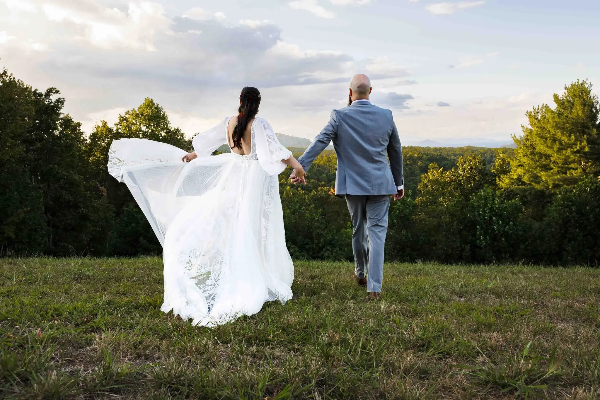 Couple walking hand in hand away from the camera toward a ridge view, her skirt swirling