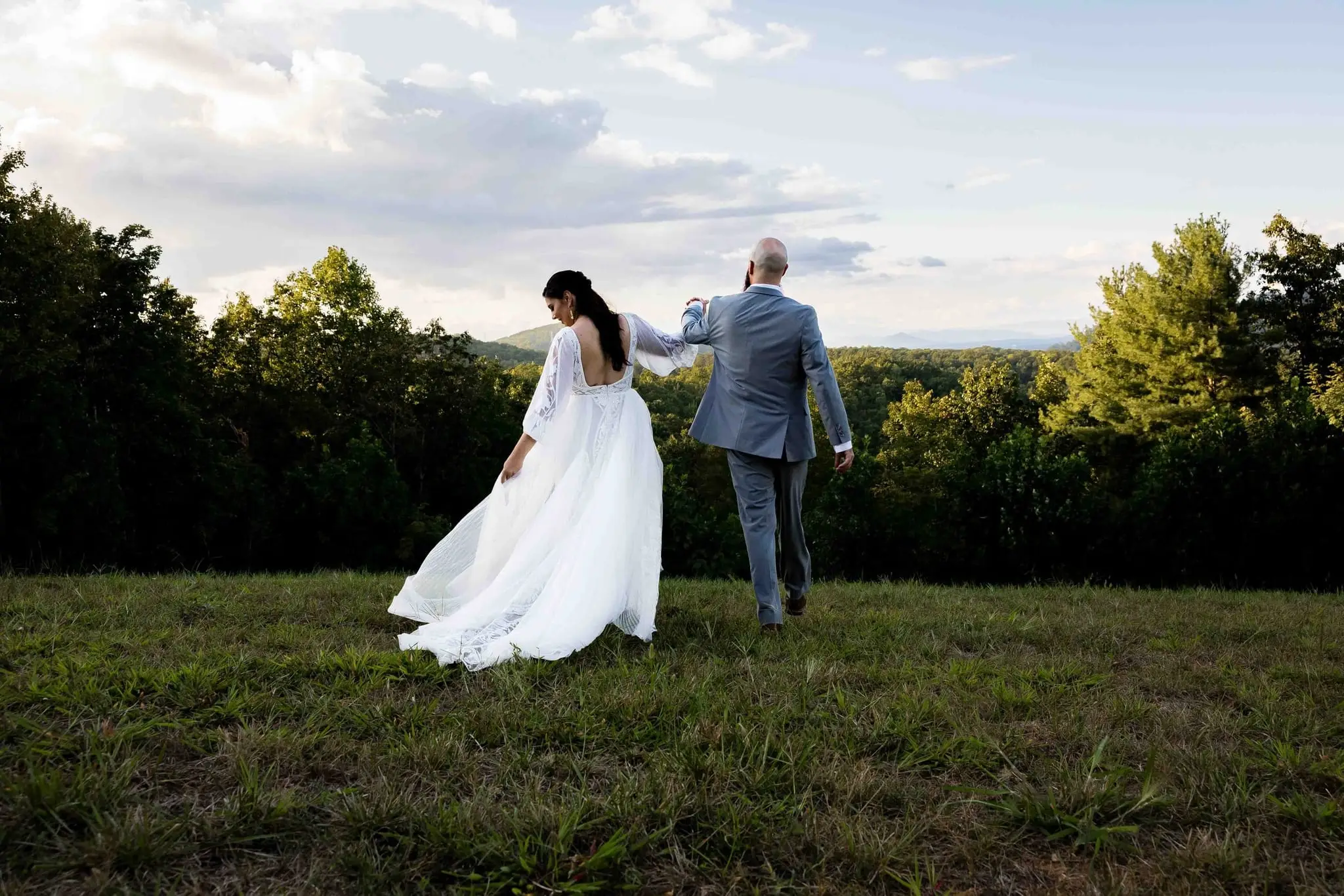 Bride and groom walking away together, her gown fluttering, toward the treeline