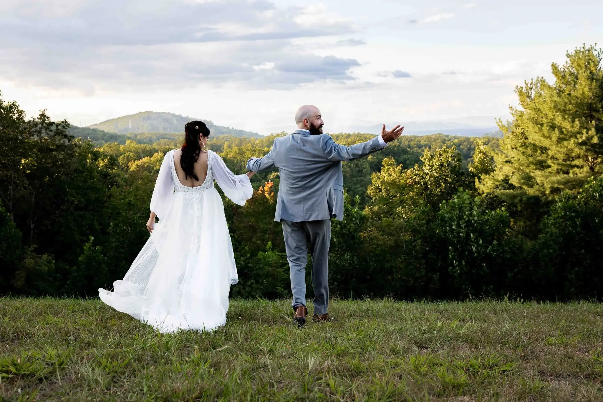 Bride and groom walk hand in hand across a grassy hilltop toward a Blue Ridge view
