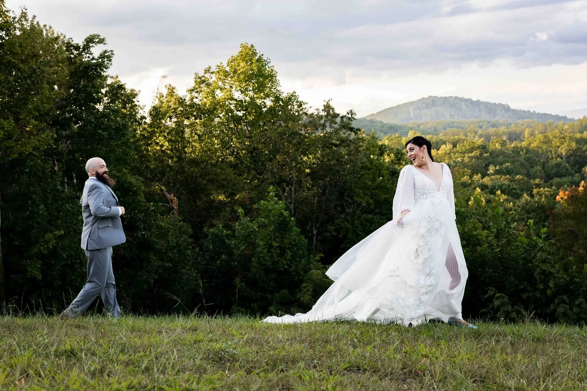 Groom walking through the frame as the bride gathers her flowing gown on the hilltop