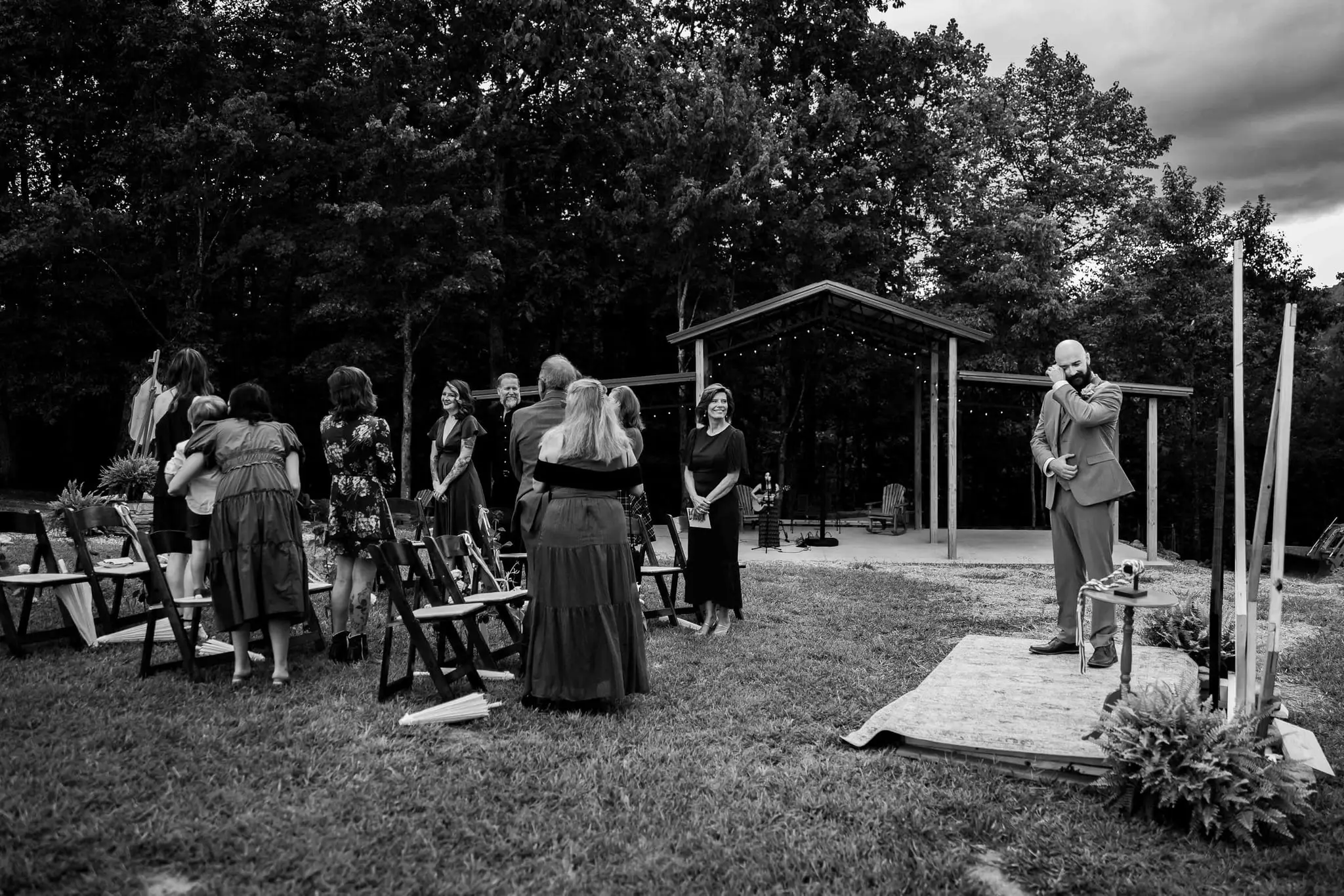 Black and white of guests gathering on the lawn near the wooden pavilion as the groom waits at the ceremony site