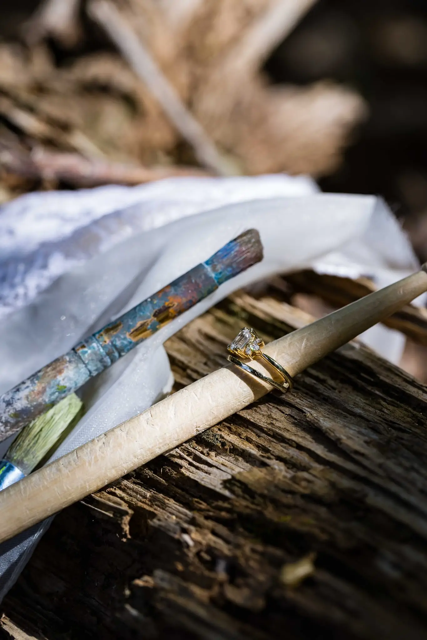 Engagement ring on a weathered log beside a paintbrush and a fold of bridal veil