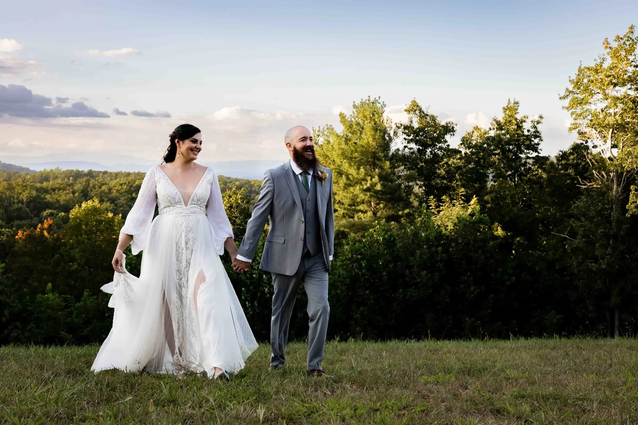 Bride and groom stroll hand in hand across the lawn with forested hills behind them