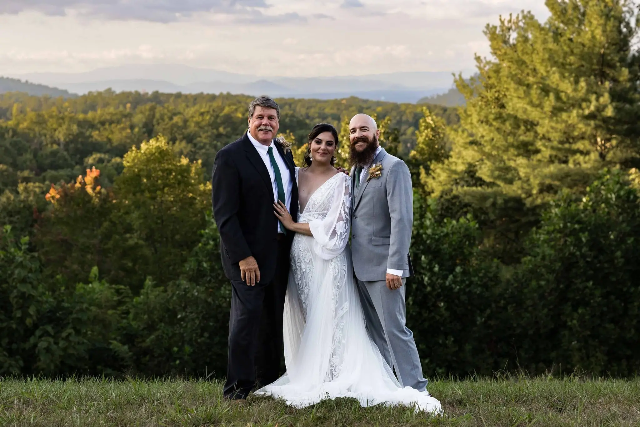 Bride and groom flanked by an older man in a dark suit on a hilltop overlooking forested hills