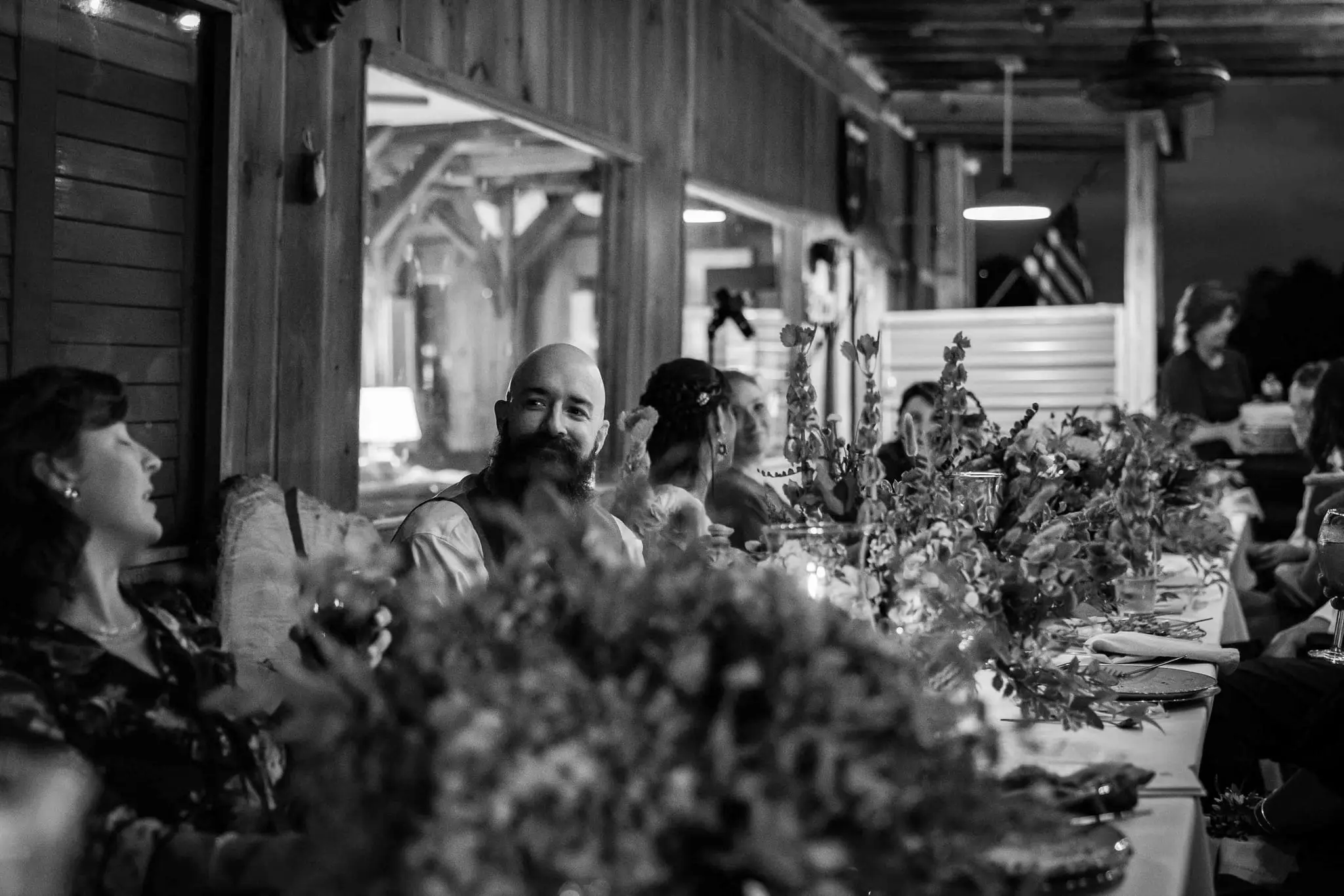 Black and white of the groom and guests laughing along a long reception table lined with greenery