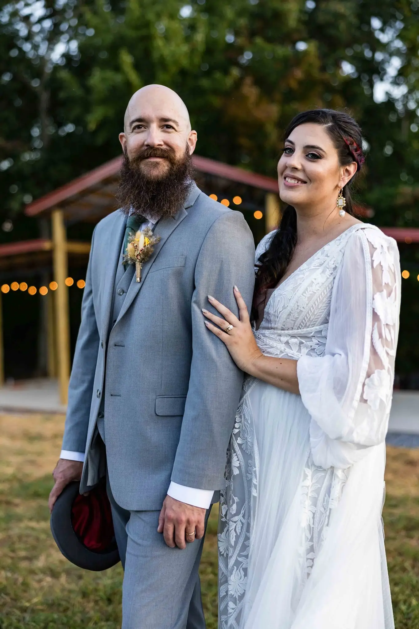 Smiling groom with his hat in hand and the bride holding his arm in front of a string-lit pavilion at dusk