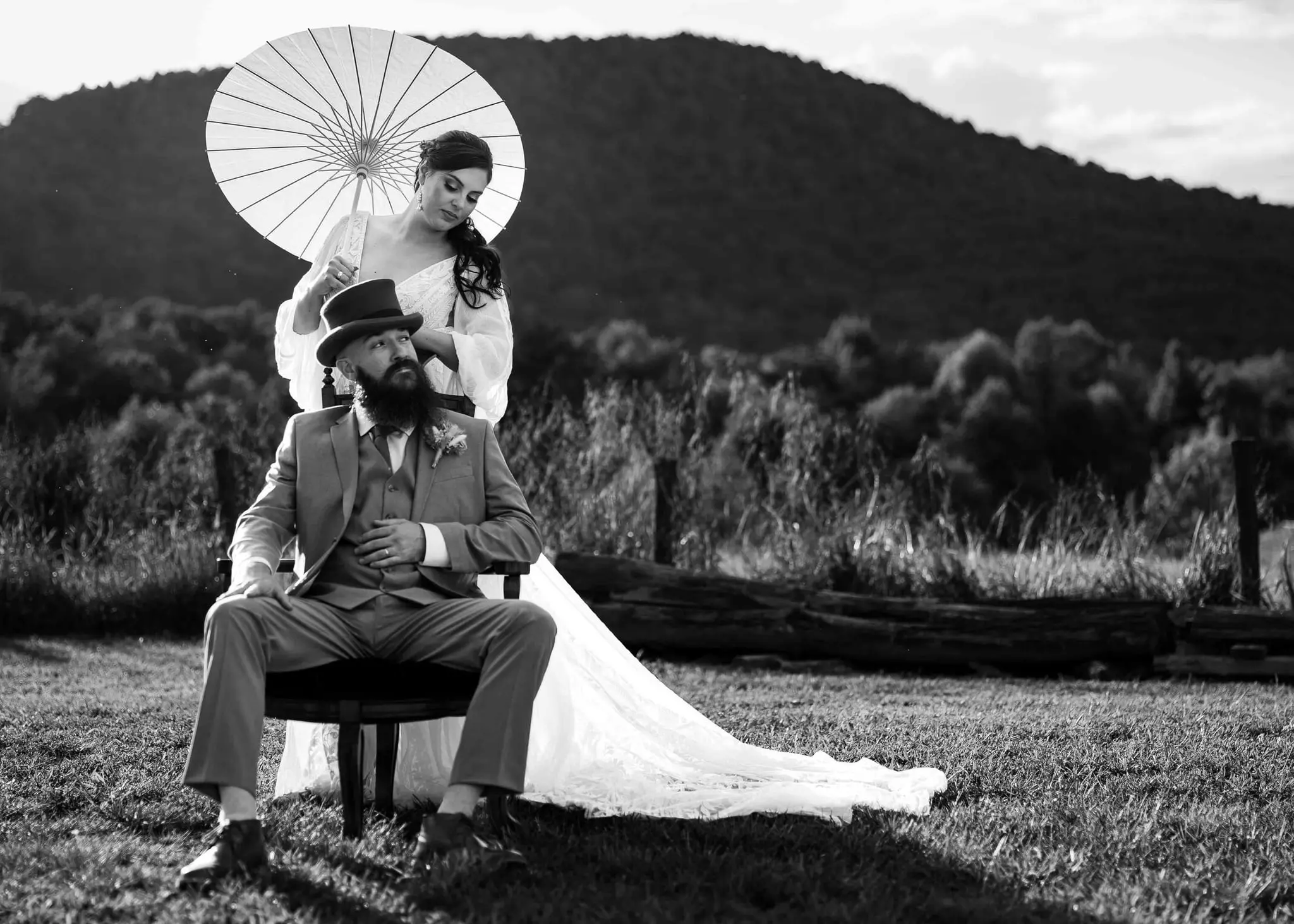 Black and white of the groom seated in a chair as the bride stands behind him holding a paper parasol