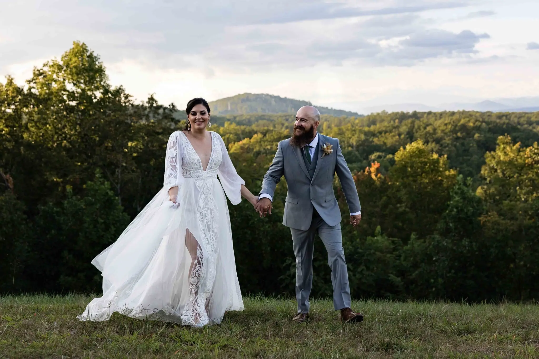 Couple holding hands on the hilltop lawn as her skirt sways, layered ridges in the distance