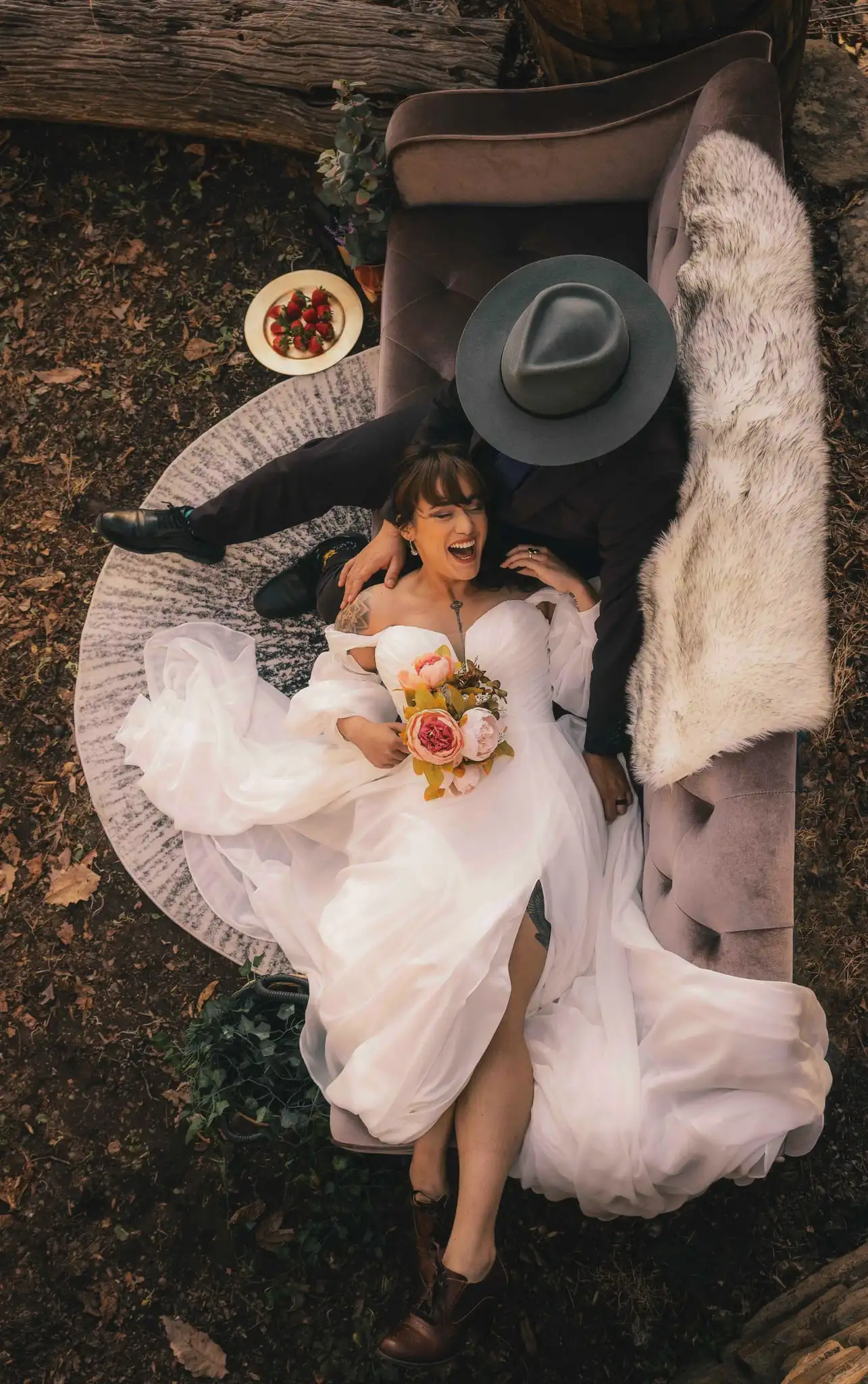 Overhead shot of a laughing bride holding a bouquet on a velvet settee, the groom's hat resting nearby