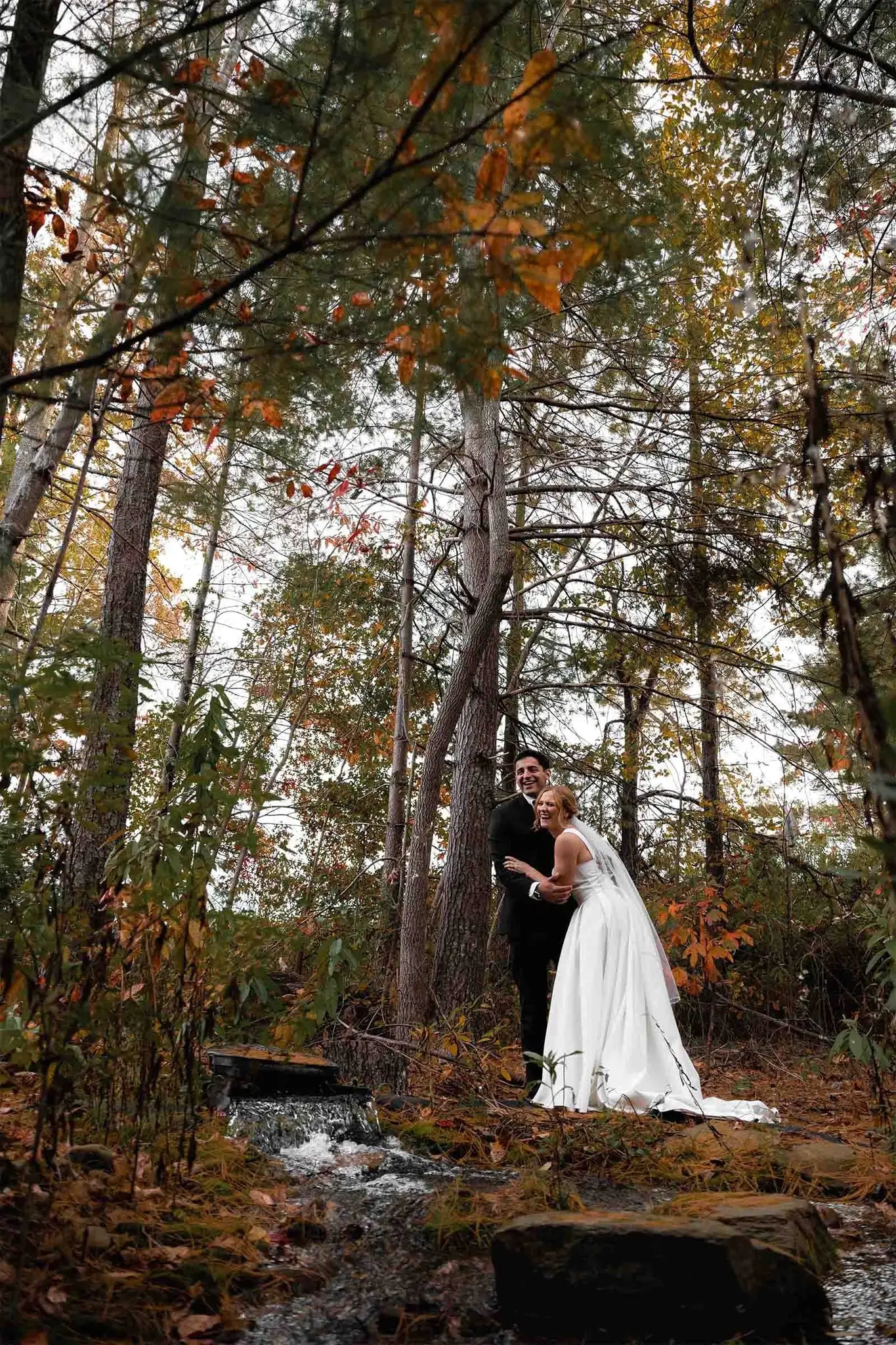 Couple in wedding attire embracing in an autumn wooded clearing beside a small stream