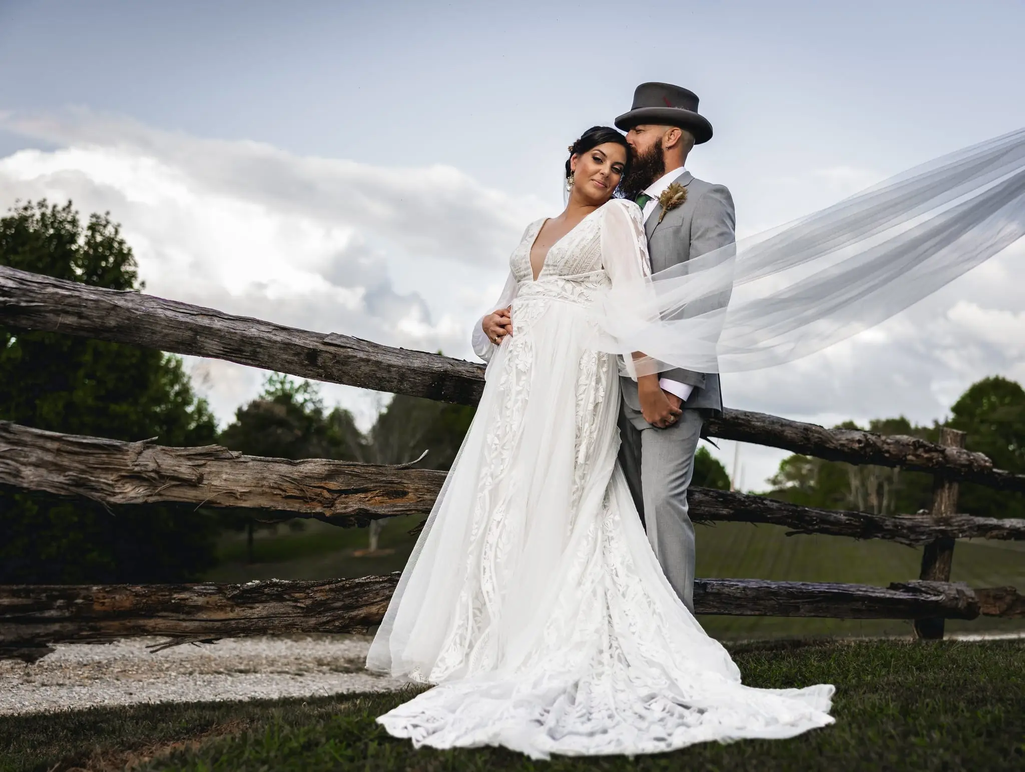 Bride and groom beside a split-rail fence as her long veil streams in the wind
