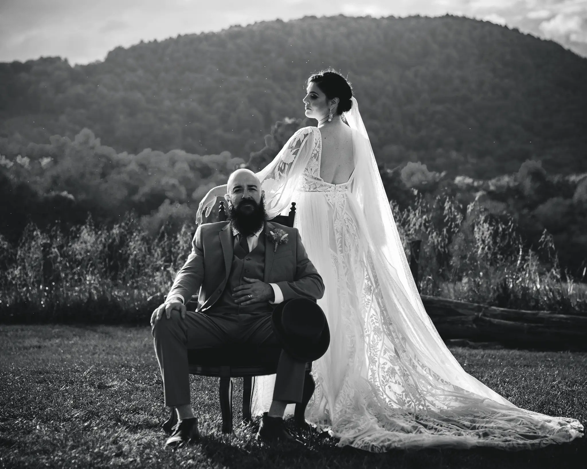 Black and white of the groom in a vintage chair as the bride stands in her long veil before a mountain