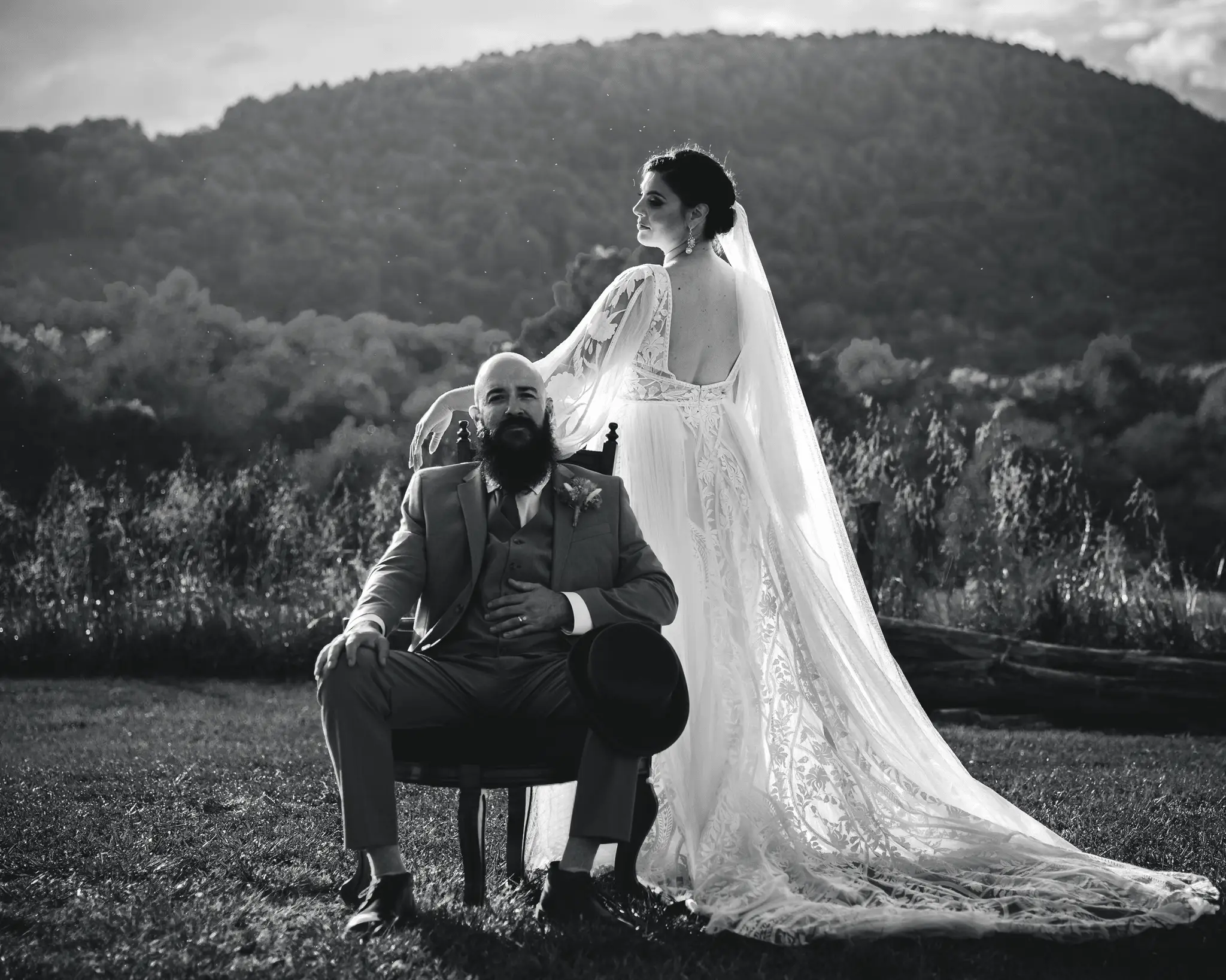 Black-and-white portrait of a bride standing behind her seated groom with mountains behind them