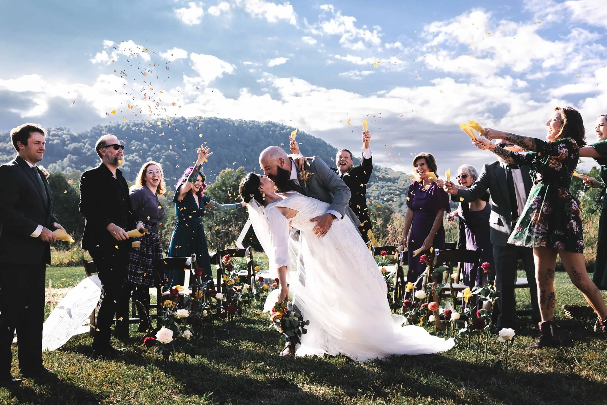 Groom dipping his bride for a kiss as guests throw confetti in a mountain meadow under a bright sky
